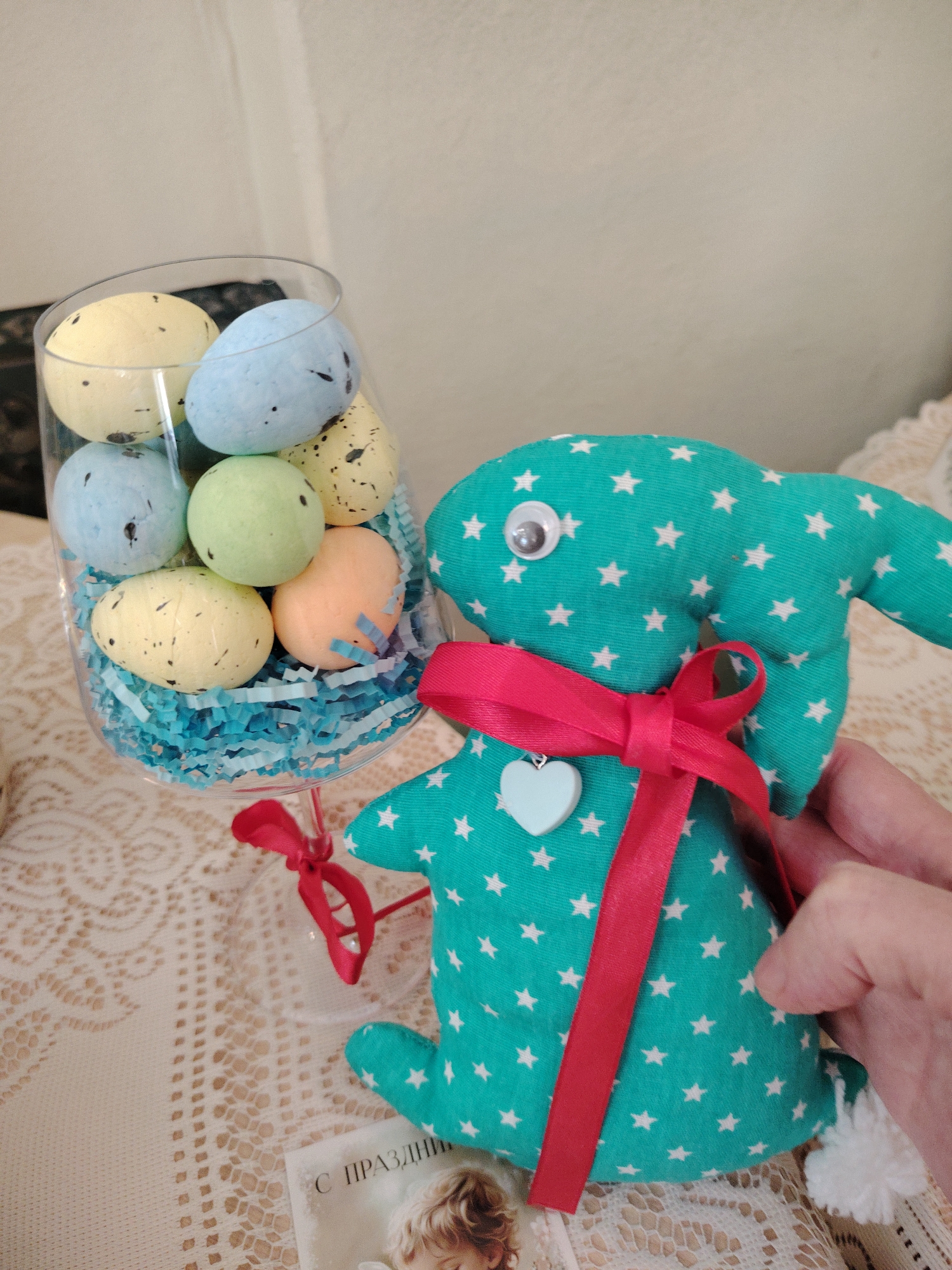 A close-up shot shows Easter decorations, likely indoors. On a lace-covered surface sits a clear wine glass filled with blue shredded paper and speckled Easter eggs in various pastel colors (yellow, blue, green, and orange). To the right, a handcrafted green fabric Easter bunny with white stars, a googly eye, a white heart pendant, and a red ribbon. A hand holds the bunny. The edge of a greeting card with Cyrillic text and an image is visible in the foreground. The location is suggested to be Sarapul, Russia, though this cannot be verified from the image alone.