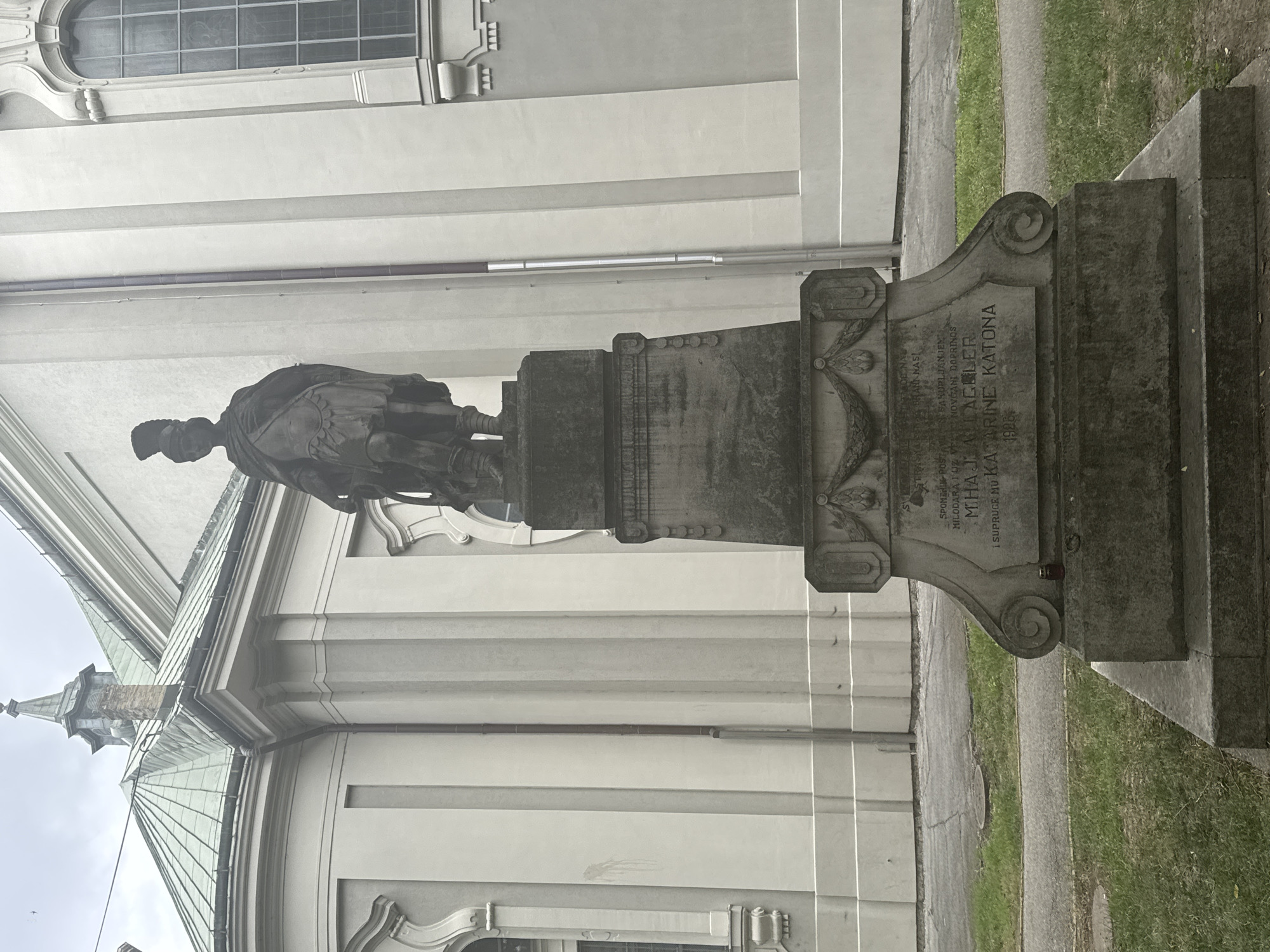 The image shows a statue of a man in Roman soldier's attire, standing on a stone pedestal in front of a church in Subotica, Serbia. The statue is made of dark stone and the pedestal is made of weathered grey stone. The church behind the statue has a green tiled roof and light-colored walls. The area around the statue is paved with grass and a pathway. There is some text inscribed on the pedestal, but it is difficult to read in the image. The statue appears to be a memorial.