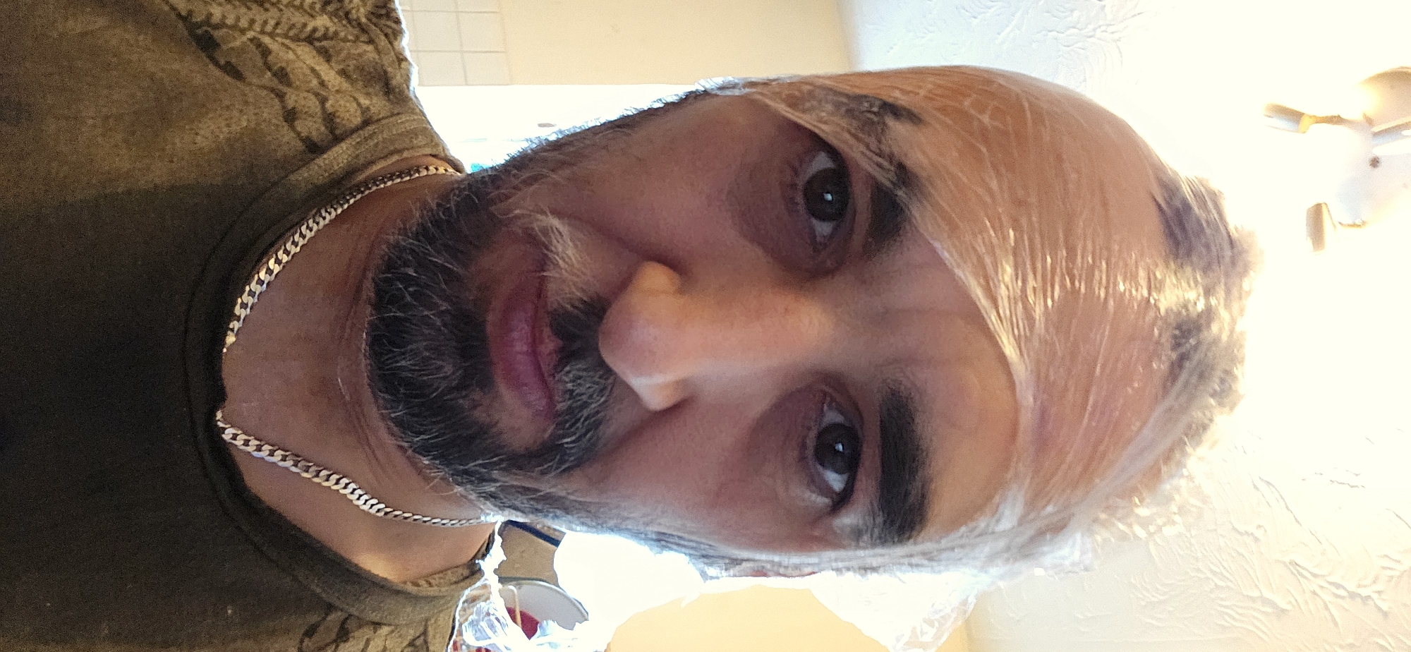 The image is a close-up portrait of a man, likely middle-aged, looking directly at the camera. He has a dark, full beard with prominent graying hairs and dark brown eyes, maintaining a neutral to slightly contemplative expression with faintly pursed lips. He is wearing a dark, possibly olive green or brown, casual top, and a silver-colored cuban link chain necklace is visible around his neck, partially obscured by his shirt collar.
A distinctive feature is the clear plastic wrap covering his entire head, suggesting he is undergoing a hair treatment such as dyeing or conditioning. Dark patches are visible beneath the plastic wrap, indicating the application of a product to his hair or scalp.
The setting appears to be indoors, likely a home environment, with bright overhead lighting creating a slight glare on the plastic wrap and illuminating his face. Parts of a ceiling and wall are visible in the background. No specific location cues for Norwich, UK, are visible within the image itself, nor is there any discernible text. The time of day cannot be determined from the indoor lighting.