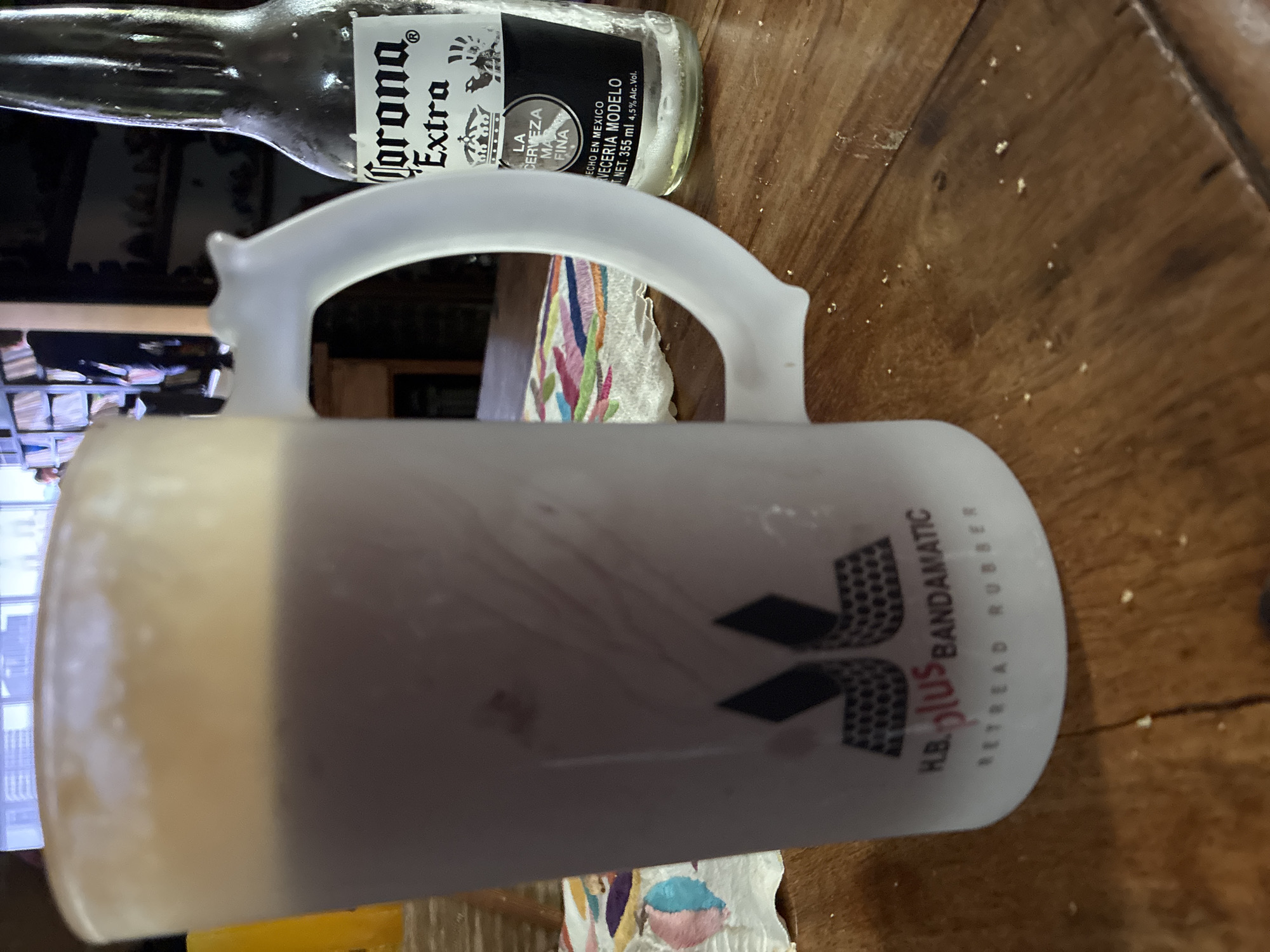 A close-up, slightly angled shot shows a frosty beer mug and a Corona Extra beer bottle on a wooden surface. The beer mug is a translucent white, with a large handle and the words "H.B. PLUS BANDAMATIC RETREAD RUBBER" visible on its side, along with a logo of some sort. It appears to be filled with a dark liquid, possibly dark beer. Partially visible behind the mug is a Corona Extra beer bottle with its label showing the beer's name and other details, including that it is made in Mexico and contains 355ml of liquid. The wooden surface appears to have slight wear and tear with some scattered particles. A colorful cloth or napkin is also visible beneath the mug and bottle. The setting does not provide enough detail to determine the location in Cañada de Madero, Mexico.