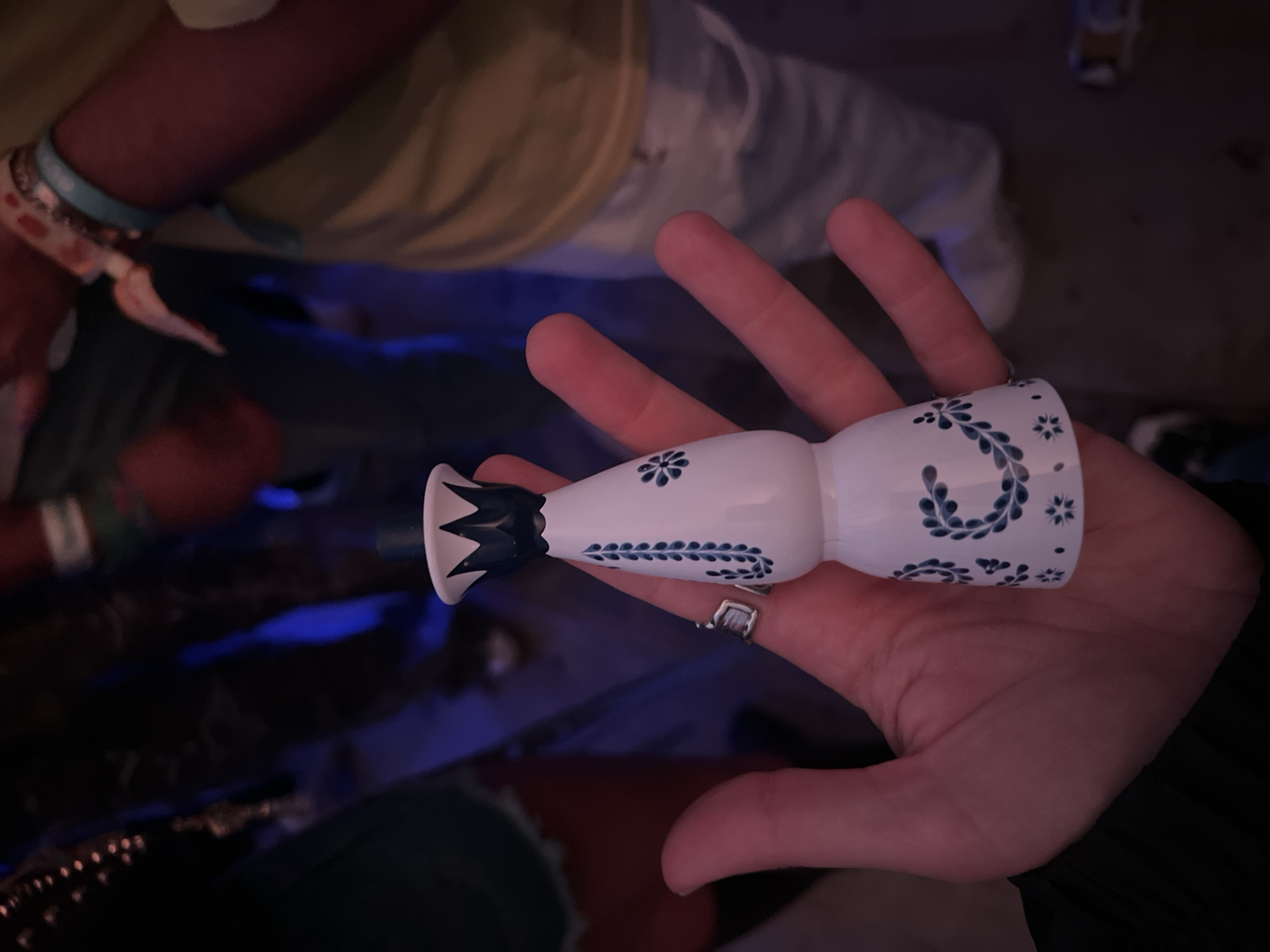 The image shows a white ceramic object with blue floral patterns held in a person's hand. The object is shaped like a vase or pipe, with a wider body and a narrower neck that flares out at the end. The location is Indio, United States of America, based on information provided. The background is dimly lit and blurry, suggesting an indoor setting. The person holding the object is wearing a ring. Other people and objects are visible in the background but are out of focus.