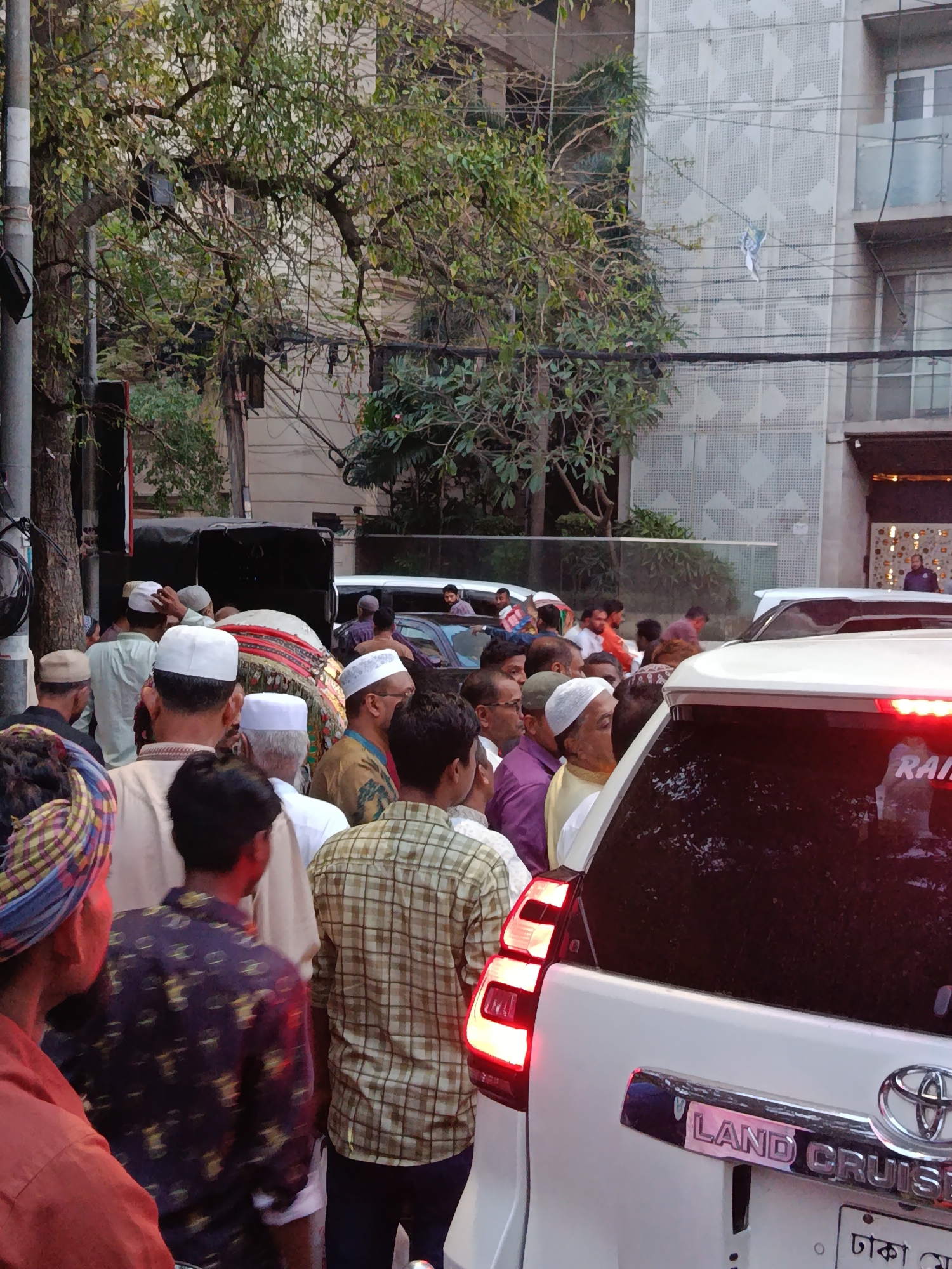 The image shows a crowded street scene in Dhaka, Bangladesh. A large group of men, many wearing Muslim skullcaps, are gathered around a vehicle with speakers, suggesting a public gathering or procession. A white Land Cruiser is parked nearby, with its taillights illuminated. The backdrop includes buildings, trees, and utility poles typical of an urban environment.