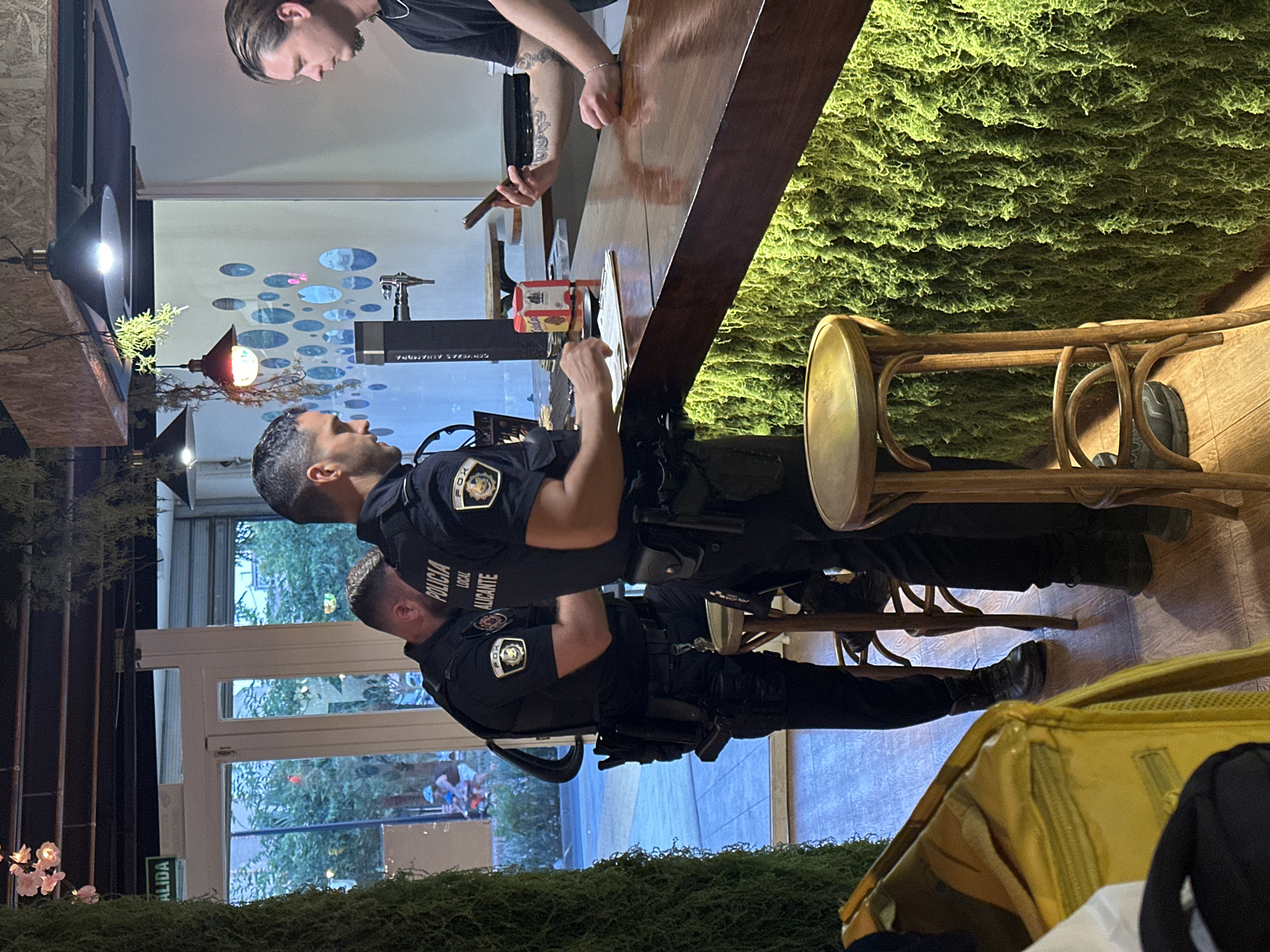 Two police officers in uniform are present at a bar in Alicante, Spain. The officer on the left, facing away from the camera, is wearing a black uniform with "POLICIA LOCAL ALICANTE" visible on his sleeve. He is standing and appears to be speaking with the officer on the right. The officer on the right, also in uniform, is facing the camera and holding a smartphone. A third individual, a civilian, is on the other side of the bar, holding his own smartphone. The bar counter is made of dark wood and is decorated with a vibrant green moss-like material. In the background, a window shows an outdoor scene with trees and people. The overall atmosphere suggests a routine interaction between law enforcement and civilians within a commercial establishment.