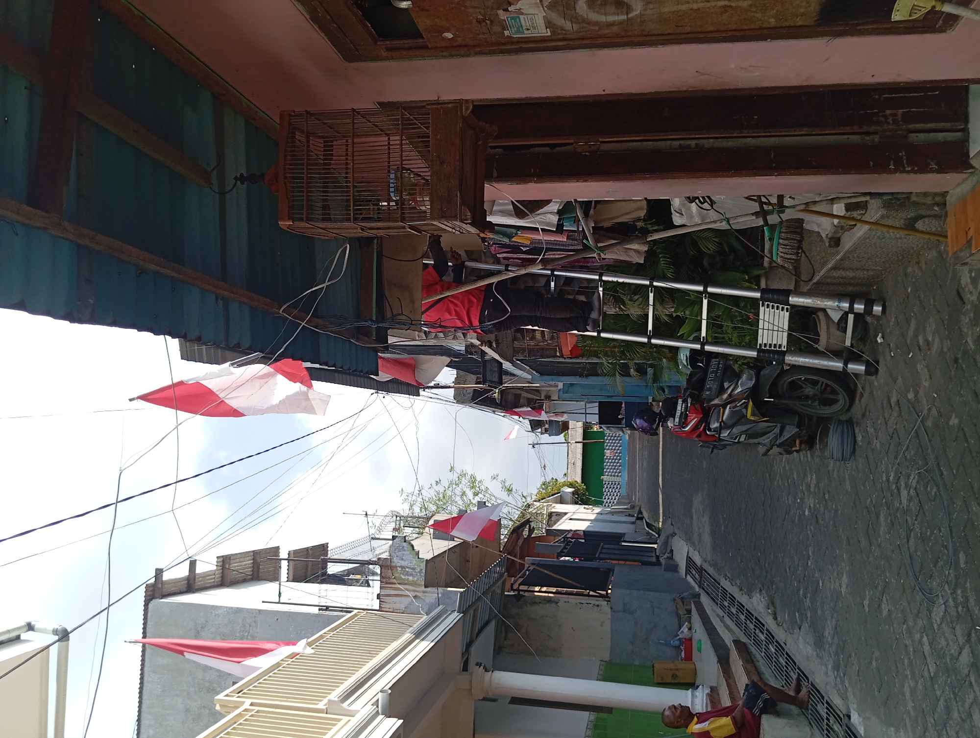 The image depicts a narrow street in Nogosari, Indonesia, lined with closely situated buildings. The scene is dominated by the presence of numerous power lines crisscrossing overhead, interspersed with several red and white Indonesian flags, suggesting a patriotic or festive occasion.
In the foreground, a man is seated on steps to the left, facing away from the camera, wearing a yellow and red shirt and dark shorts. A motorcycle is parked on the right side of the street.
Further down the street, a person in a red shirt and dark pants is on a silver, multi-section ladder, appearing to be working near a wooden birdcage suspended from an overhead structure. Clothes are hanging on a line nearby. The buildings on either side of the street show signs of wear, with some having balconies or extended roofs. The street itself is paved with textured grey stones. The overall impression is of a residential area with a dense, urban atmosphere.
