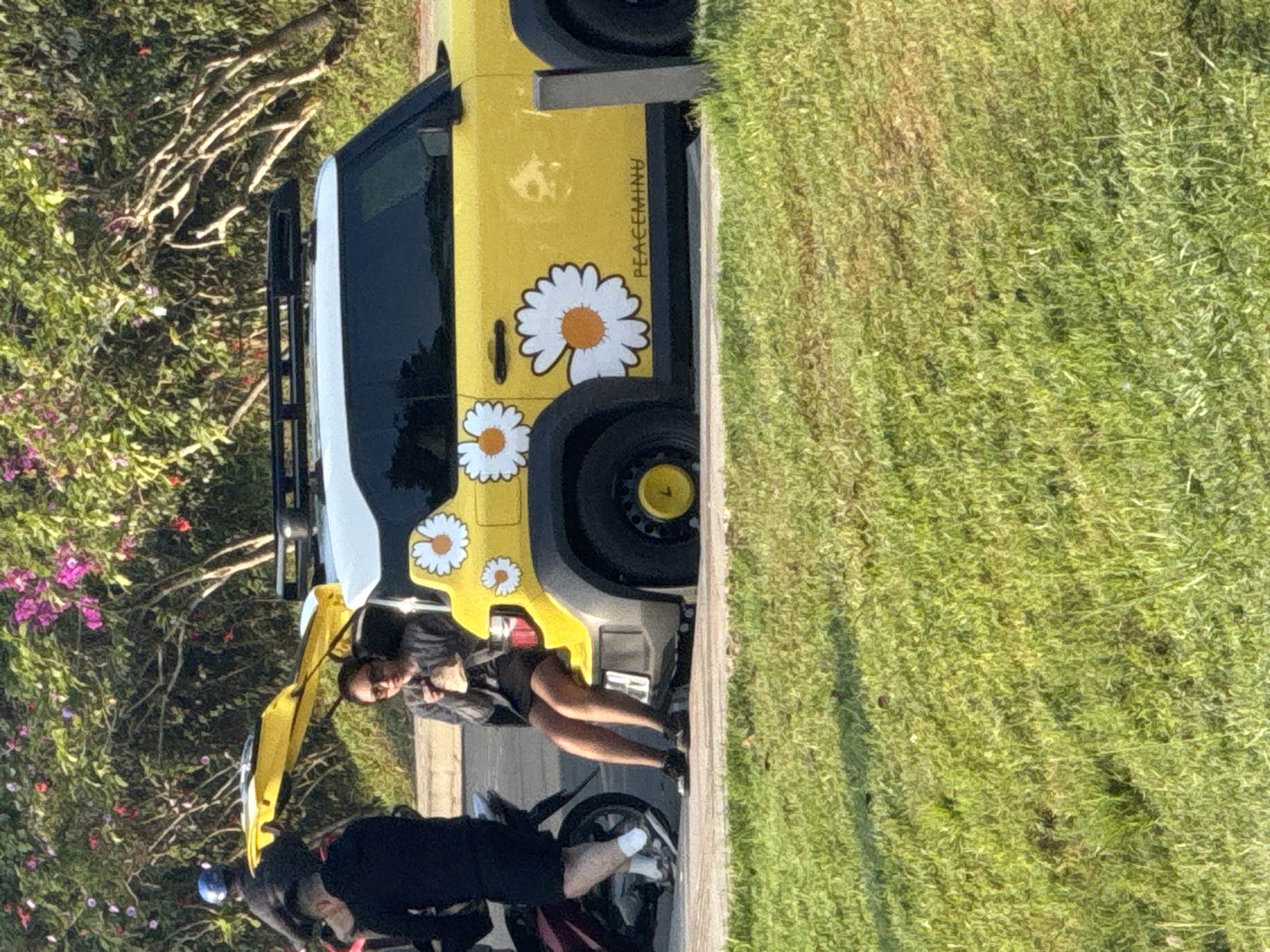 The scene captures a yellow and white SUV decorated with daisy decals, parked alongside a grassy area near possibly Xuan Huong Lake in Da Lat, Vietnam. Two individuals are visible near the vehicle. One is female, clad in dark clothing and sunglasses, and leans against the car's fender. The other is male, dressed in dark attire with white socks and shoes, near a motorcycle with a red detail. The background features foliage and some trees and flowers.