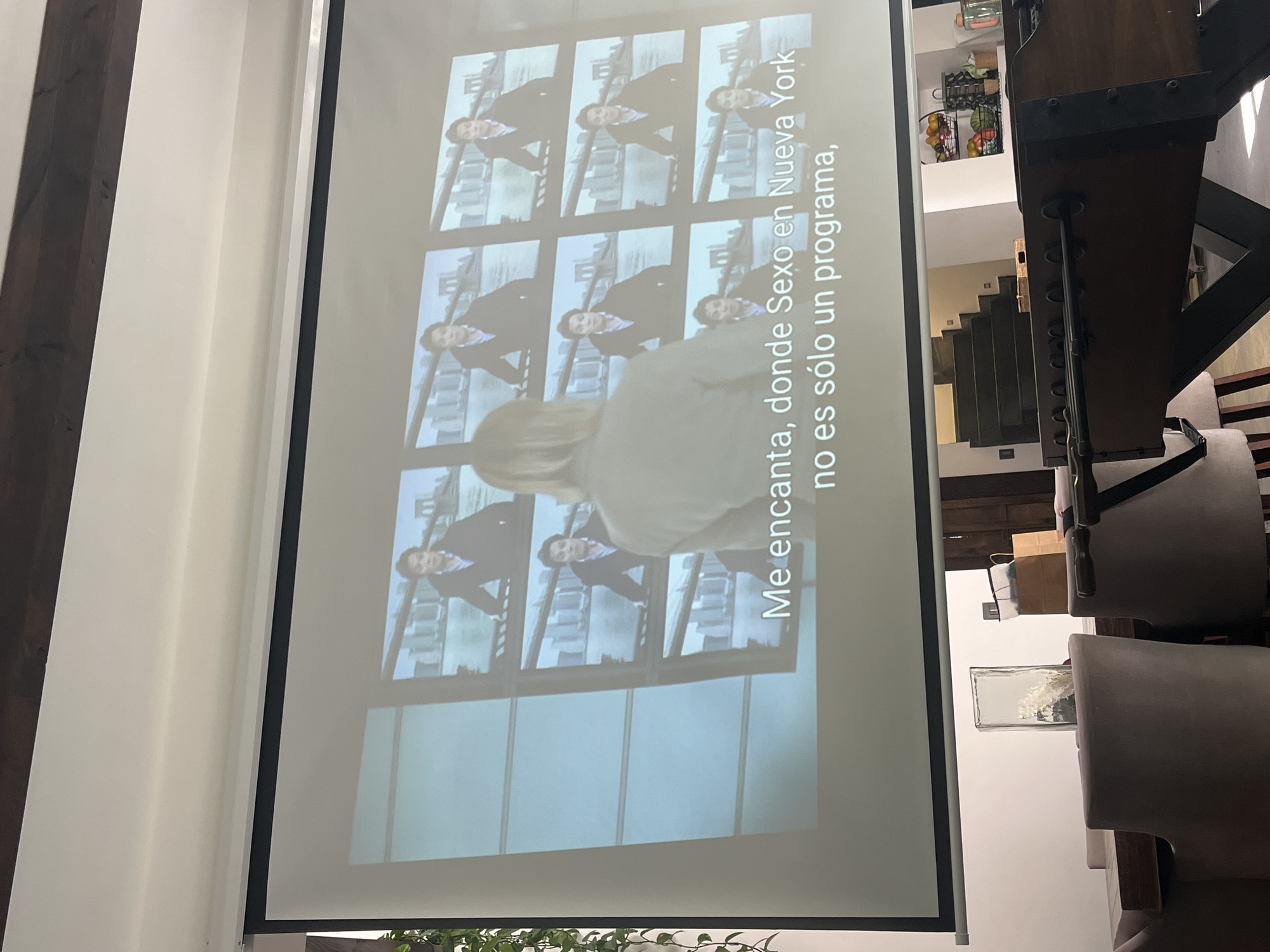 A projected image shows a person watching "Sex and the City" on a screen in San Bernardino Tlaxcalancingo, Mexico. The screen displays a grid of images featuring a man in a suit. Subtitles in Spanish read, "Me encanta, donde Sexo en Nueva York no es sólo un programa," indicating the person enjoys the show. The image is taken indoors with a partially visible dining table, chairs, stairs, and a shelf with items.
