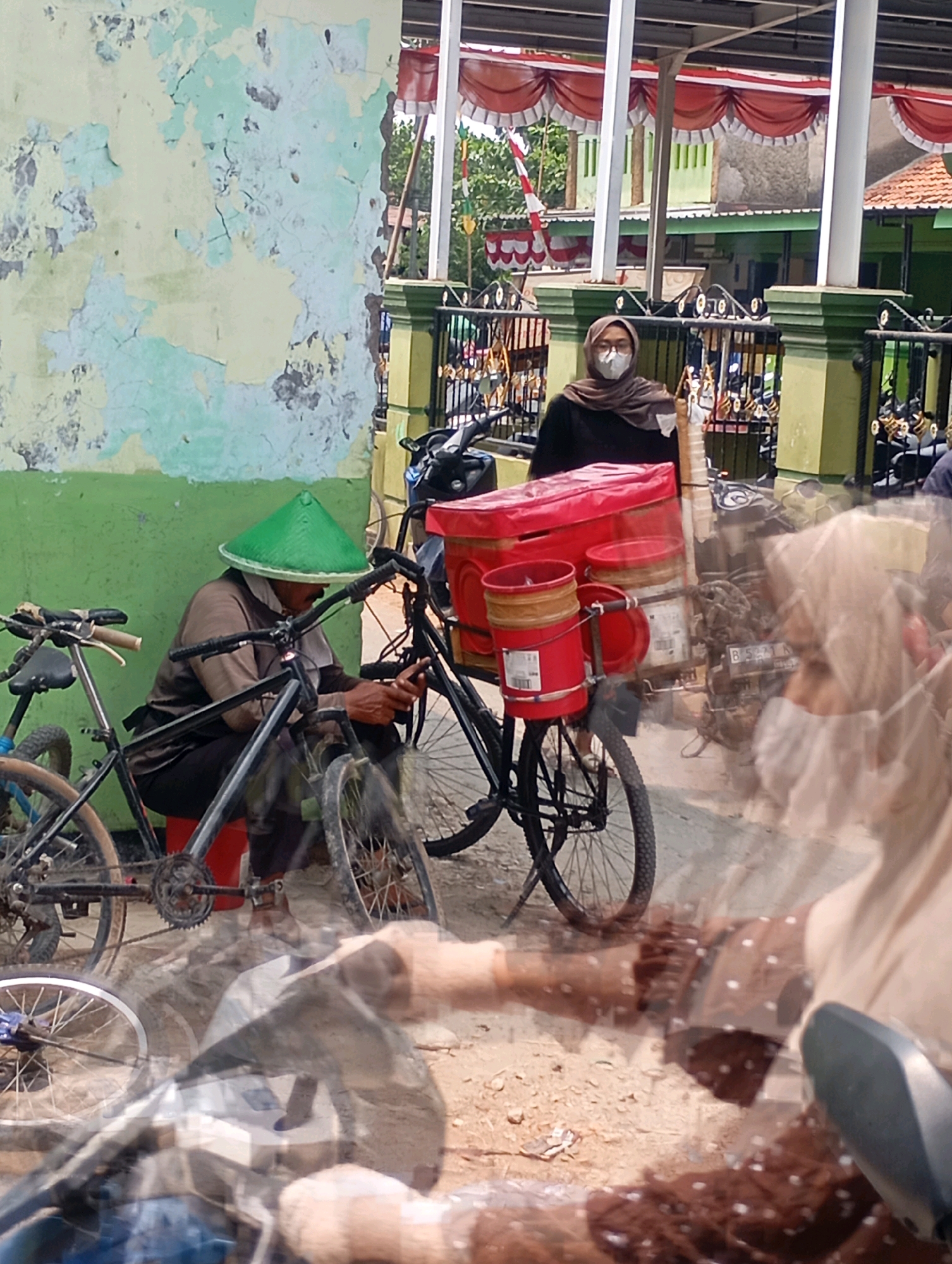 A person wearing a green conical hat sits on a red stool next to a bicycle, with their hands on the handlebars. The bicycle has a red cargo container attached to the back. In the background, another person wearing a hijab and mask stands near a building with red and white awnings. Several motorcycles are parked behind a fence. The scene appears to be outdoors, possibly on a street or market area, with a weathered wall on the left and a building with green trim in the background. The image has a slightly distorted or blurry effect, possibly due to motion or a glass surface.