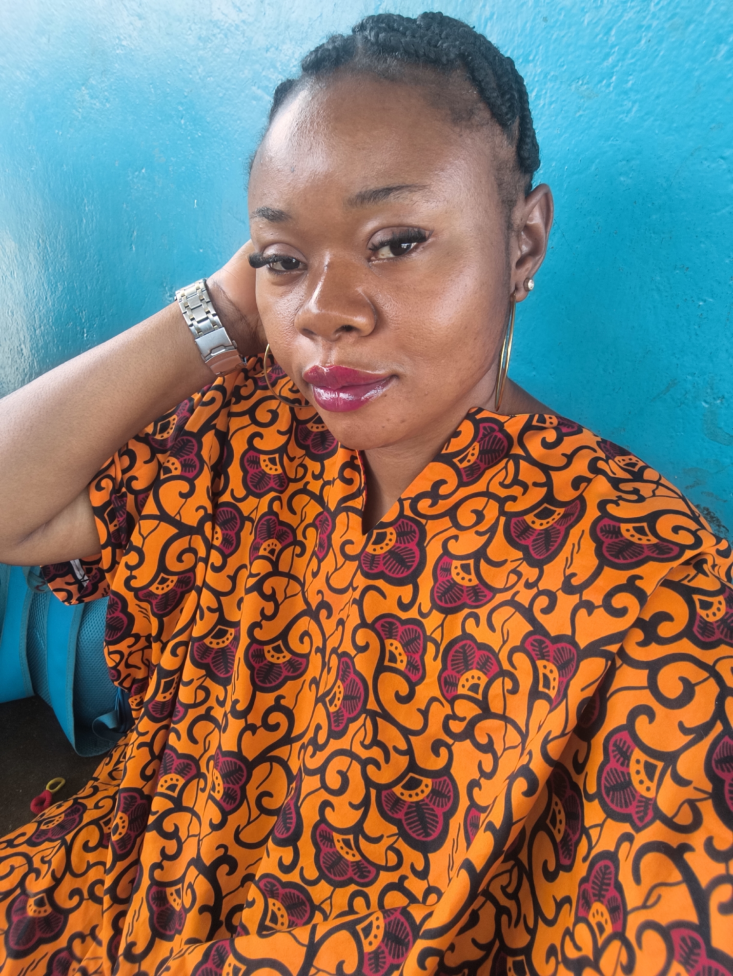 A medium close-up portrait features a dark-skinned woman with braided hair, positioned against a vibrant turquoise-blue wall. The woman, likely in her 20s or 30s, looks directly towards the camera with a composed expression. She is wearing a brightly patterned garment, possibly a kaftan or dress, made of a fabric with dominant orange and black hues, accented with magenta floral and swirling designs.
Her left arm is raised, with her hand gently resting on the back of her neck, showcasing a silver-toned metallic watch on her wrist. She wears large gold hoop earrings in both ears, with an additional small stud visible in her right earlobe. Her makeup includes long, defined eyelashes and glossy, bright magenta lipstick.
The blue background appears textured, suggesting a painted wall. To the lower left, a portion of a blue backpack or bag is visible, partially obscured by her clothing. The lighting is bright and even, indicating a daytime setting. The woman's attire and features are consistent with the provided context of Jos, Nigeria. There is no visible text in the image. The overall impression is a casual yet stylish portrait.