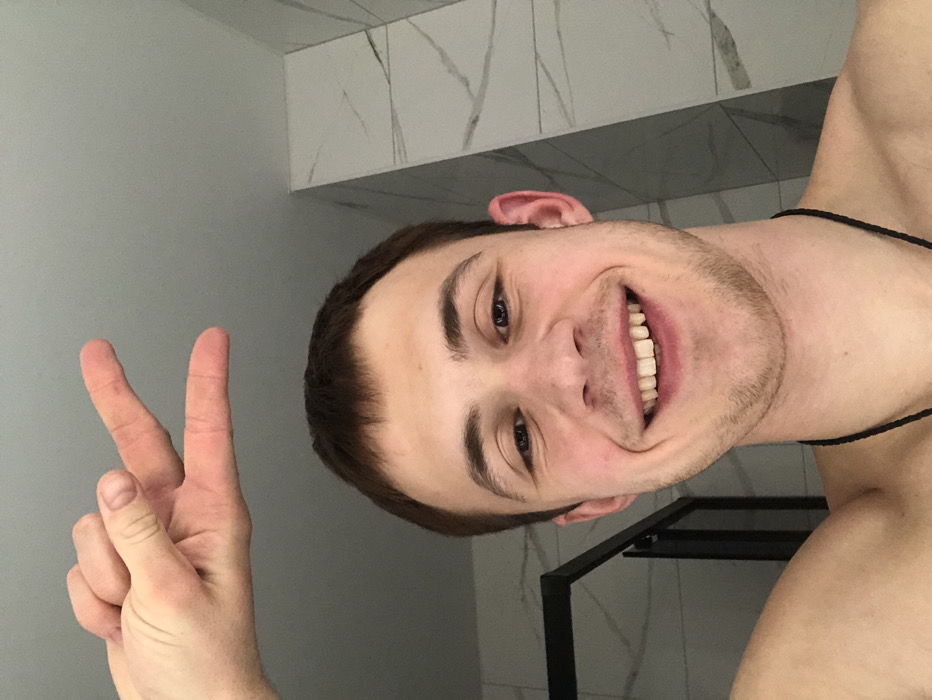 A smiling fair-skinned man faces the camera, giving a peace sign with his left hand. He has short brown hair, light stubble, and brown eyes. He appears shirtless, wearing a thin black necklace. The background features light gray and white marbled tile, and a dark metal structure is partially visible behind him. The image is rotated counter-clockwise.