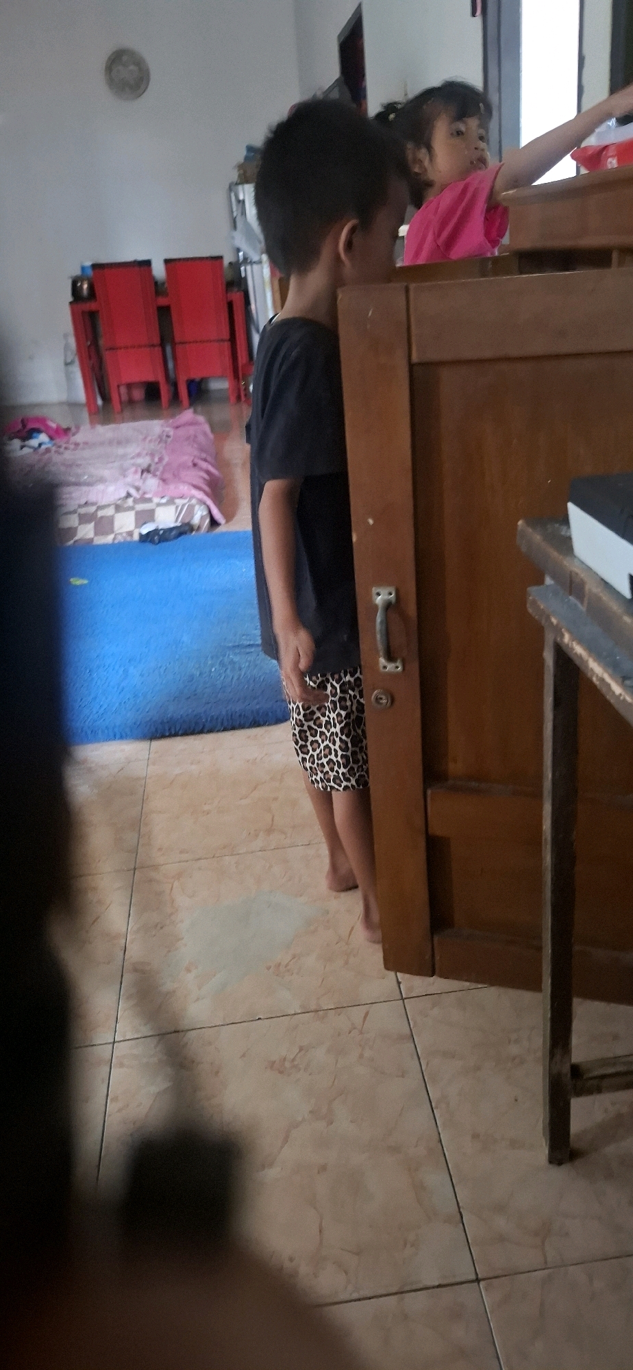 The image captures a scene inside a home in Cibinong Hilir, Indonesia. A young boy wearing a black t-shirt and leopard-print shorts stands partially obscured by a wooden cabinet. He appears to be looking towards the right side of the frame, away from the camera. Behind him and to the right, a young girl in a pink t-shirt is visible, reaching out her arm. In the background, red chairs and a mattress with bedding are visible, suggesting a living or sleeping area. The floor is tiled with a light-colored, patterned material. The lighting appears to be natural, coming from an unseen source. The overall impression is of an informal, domestic setting with children interacting or playing.