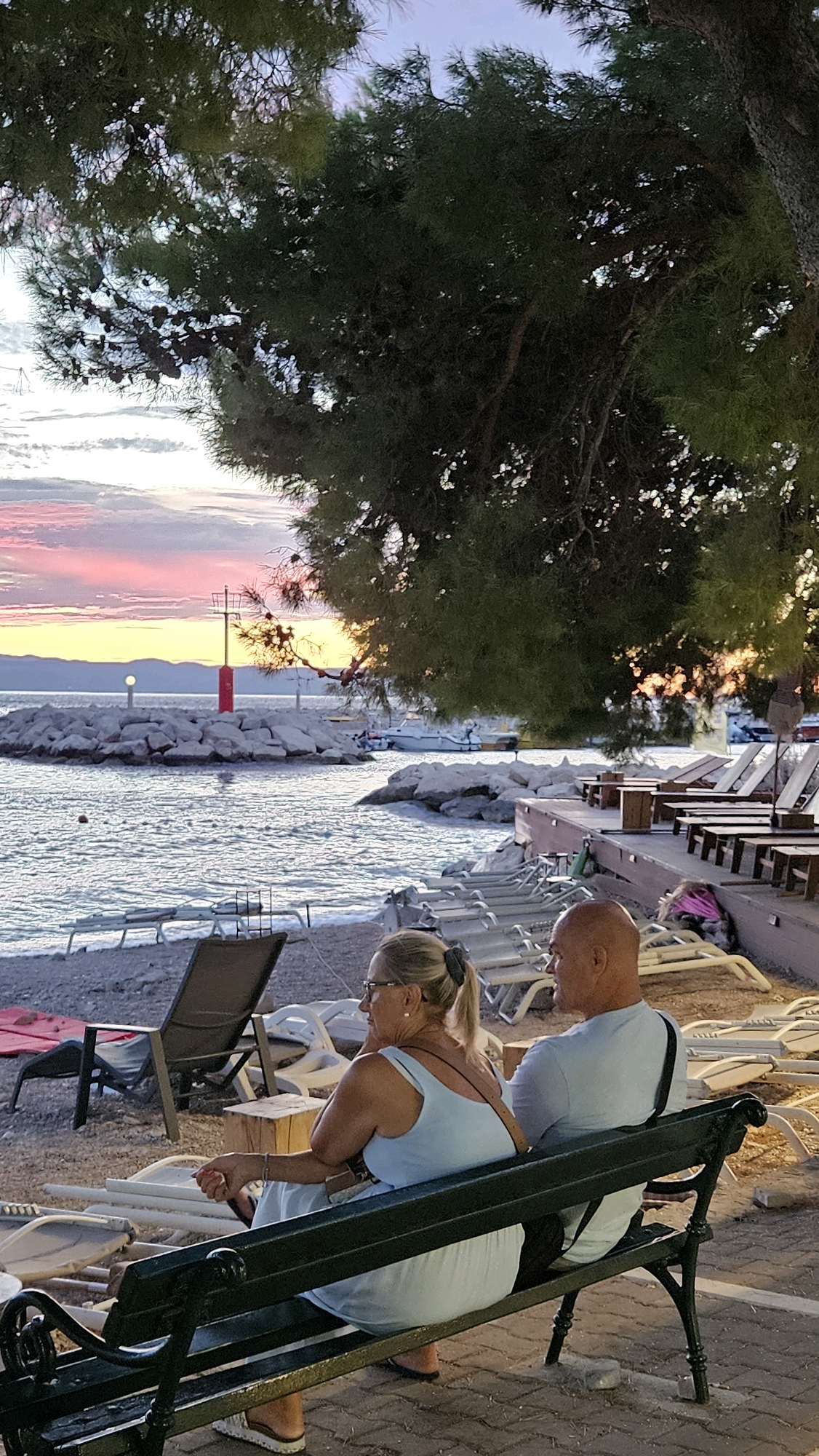 The image depicts a couple seated on a dark green bench, facing right, with their backs partially towards the viewer. They are positioned on a paved area next to a pebble beach. The woman, on the left, wears a light blue sleeveless dress and glasses, with her blonde hair tied in a ponytail. She is looking to her left. The man, on the right, has a shaved head and wears a light blue t-shirt. He is looking towards the right. Behind them, the beach is lined with rows of empty lounge chairs and some tables, suggesting a coastal resort area. The sea is visible, with calm water and a rocky breakwater in the distance. On the breakwater, there is a red navigation beacon. The sky displays a sunset with soft pink and yellow hues. Lush green trees overhang the scene from the upper right. The overall atmosphere suggests a peaceful evening setting in a coastal town.
