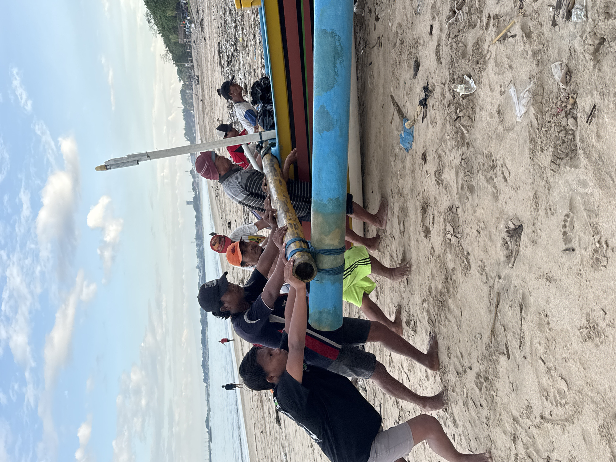A group of seven men stand on a sandy beach and appear to be lifting or moving a colorful boat. The boat is painted with yellow, black, red, and blue stripes. The men are dressed in casual clothing, including t-shirts, shorts, and hats. The beach is littered with debris. The location is likely Kuta Selatan, Indonesia, given the Indonesian background and the type of boat, which is commonly used in the area. The sky is partly cloudy. In the background is an ocean and other people.