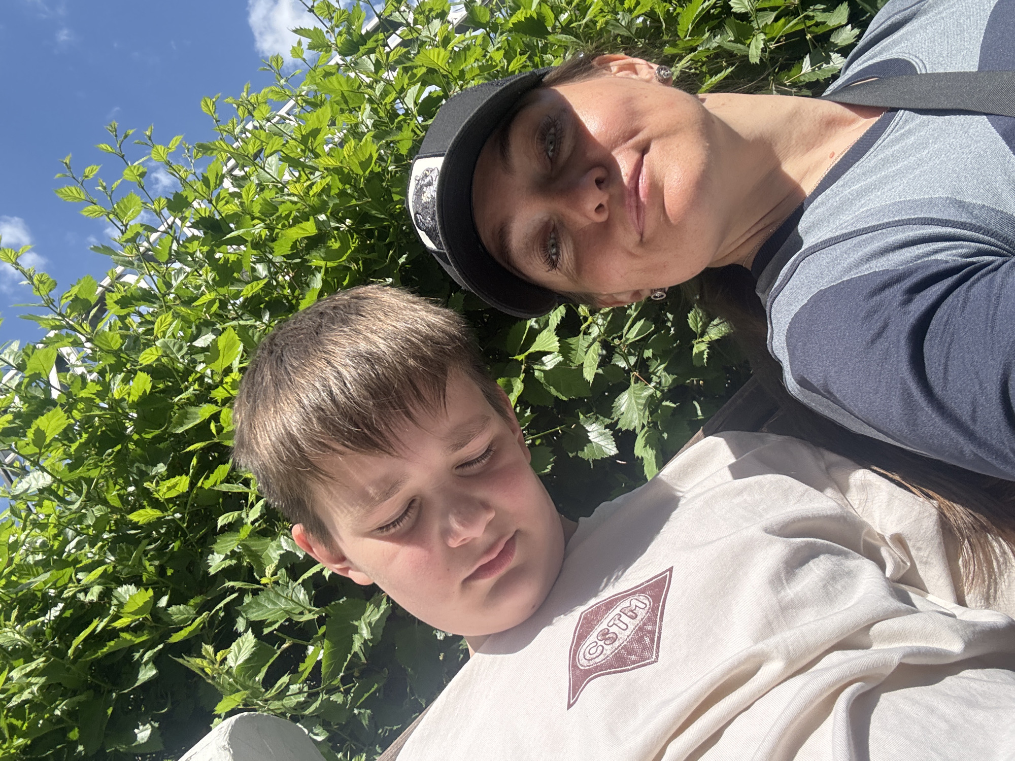 A selfie-style, eye-level photograph shows a woman and a boy against a backdrop of lush green foliage. The woman, positioned at the top of the frame, has fair skin, light eyes, and wears a black baseball cap. She is dressed in a grey and navy blue top. The boy, located towards the bottom of the image, has fair skin, brown hair, and is wearing a light beige T-shirt with a maroon emblem on the chest. The location may be Chaikovsky, Russia.