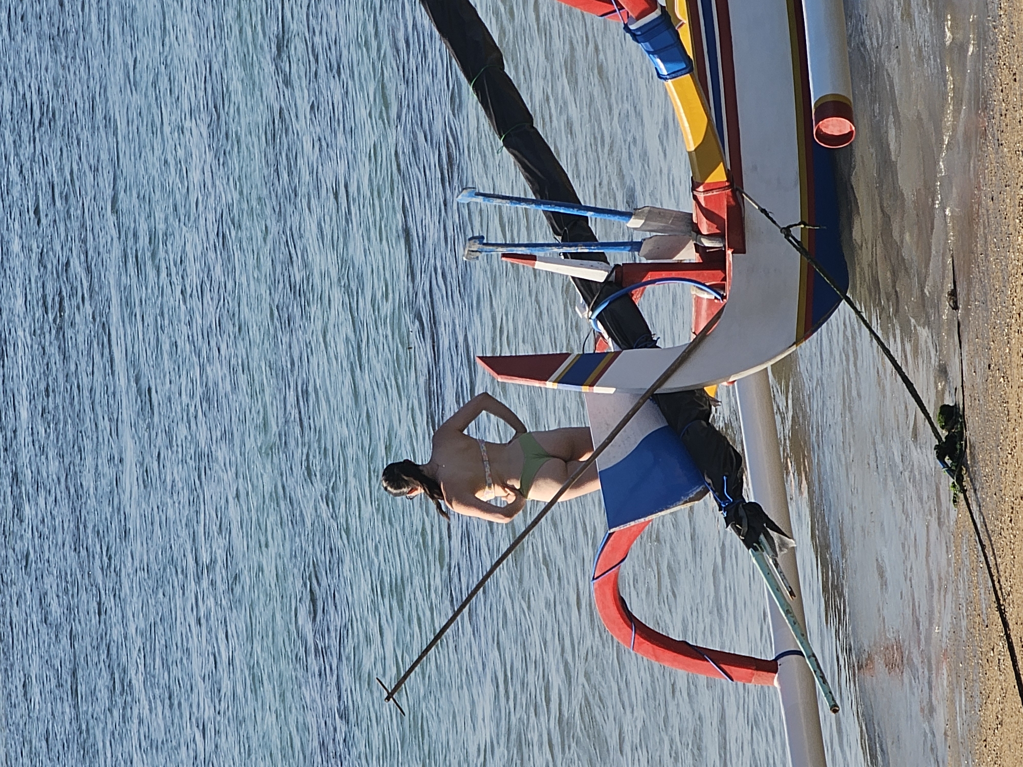 A woman in a bikini stands in the shallow water next to a colorful boat. The boat is white with red, blue, and yellow trim. Two oars are secured to the boat. The water is clear and blue, and the sand is light brown. The scene appears to be on a beach in Denpasar, Indonesia.