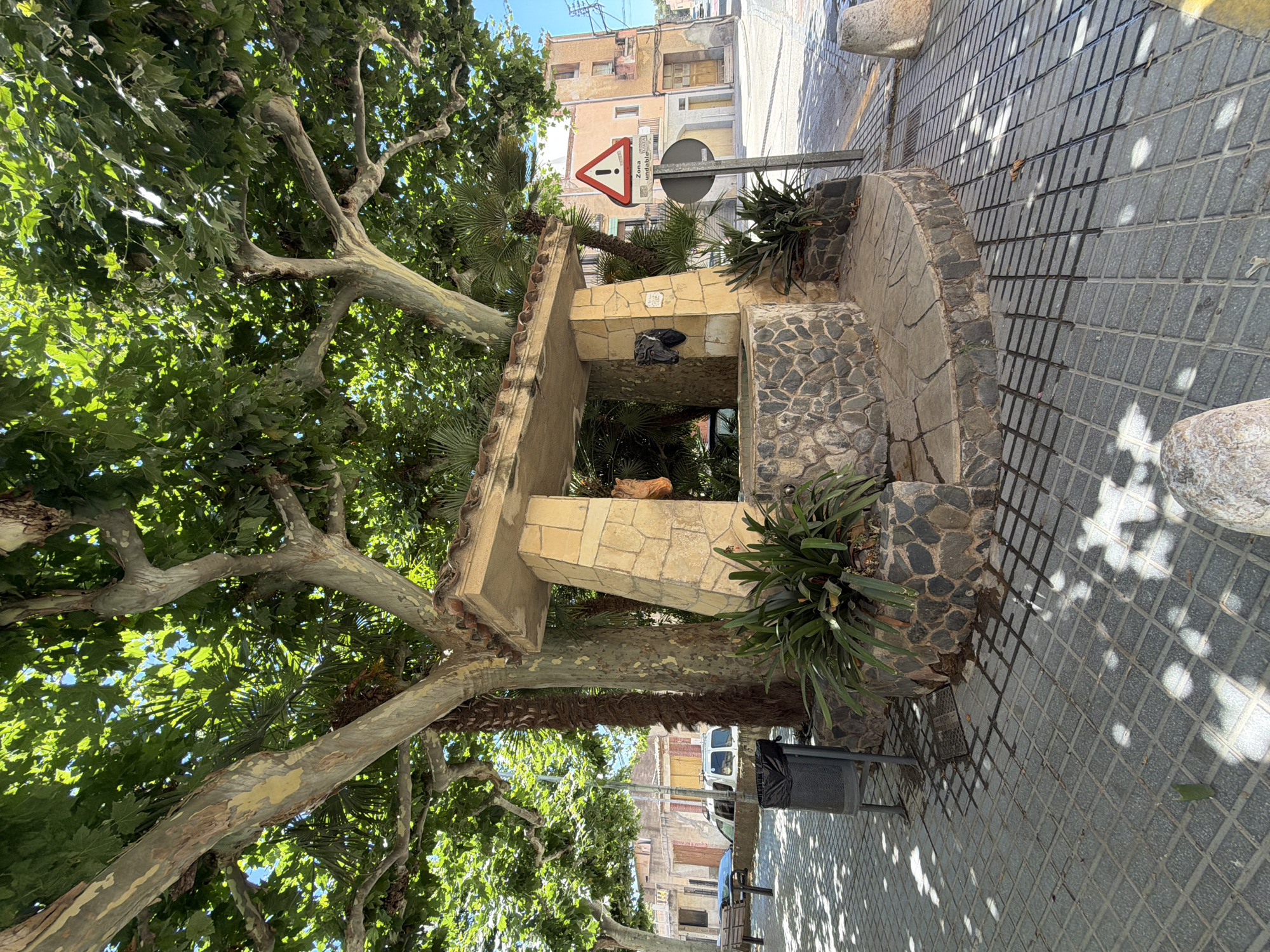 The image shows a public square in Riudoms, Spain, featuring a stone well with a tiled roof structure overhead, supported by stone pillars. The well is surrounded by a circular stone base, and lush green plants are growing around it. A large tree with broad leaves provides shade, with its branches extending over the well. In the background, there are buildings with windows and doors, typical of Spanish architecture, and a street with a traffic sign indicating a warning. A trash can is visible to the left of the well. The ground is paved with square tiles, and sunlight creates a dappled shadow pattern. There are no people visible in the image.