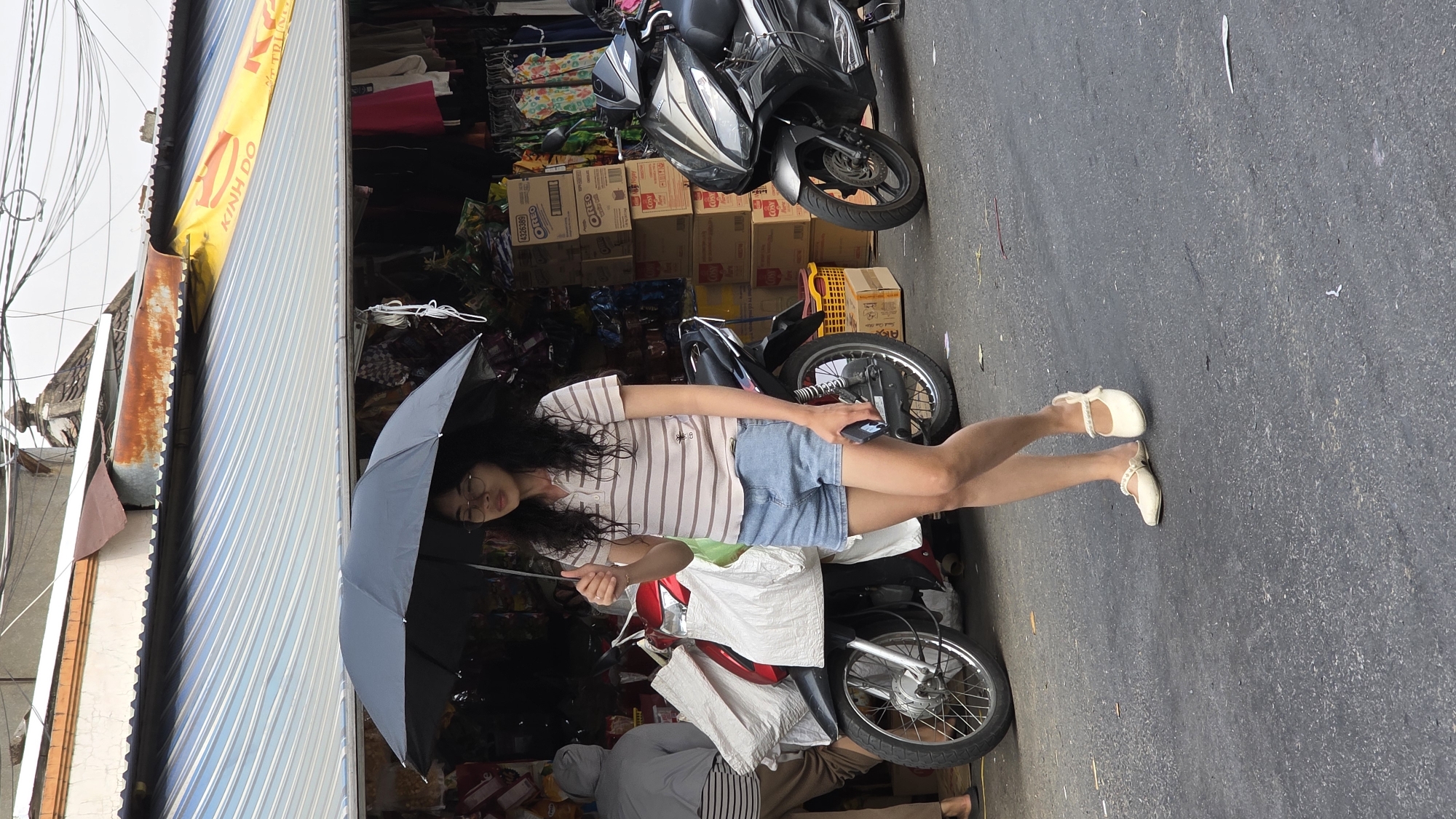 The image captures a vibrant daytime street scene in what appears to be a commercial area in Nha Trang, Vietnam.
**Subjects:** The central subject is a young woman with long, dark, wavy hair, wearing glasses, a light-colored striped short-sleeved shirt, and denim shorts. She stands beside a red and black motorcycle. Another person, partially visible in the lower left, is wearing a grey hooded top and striped pants, seemingly kneeling or seated behind the red motorcycle. Two motorcycles are parked on the asphalt street: a black scooter-style motorcycle (possibly a Honda PCX) and the red and black motorcycle (possibly a Honda Wave) next to the woman. Behind the motorcycles, the background reveals a shop or market stall filled with various merchandise, including stacks of cardboard boxes.
**Activity or event:** The woman is standing casually, holding a closed black umbrella, which could be for sun protection or carried in anticipation of rain. Her eyes are mostly closed, suggesting she might be resting, shielding her eyes from bright light, or lost in thought. The activity of the partially visible person is unclear but seems related to the shop or motorcycles.
**Notable details:** The scene is brightly lit, indicating it is daytime. The street is paved with dark asphalt, and overhead, numerous power lines are visible. The shop area features a yellow awning with red text and a blue and white striped awning. The buildings on the left show signs of age, with rusty corrugated metal and exposed utilities. The overall atmosphere suggests a typical busy urban environment in Southeast Asia.
**Visible text:** The text "KINH DO" is clearly visible in red letters on the yellow awning above the shop. The brand name "OREO" is also visible on several stacked cardboard boxes inside the shop area.