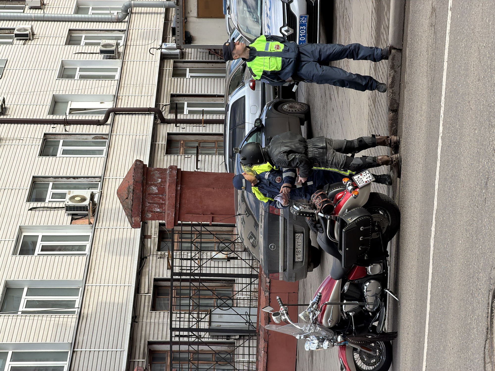 A motorcycle rider is being questioned by two traffic police officers in Krasnoyarsk, Russia. The motorcycle, red and white, is parked on the street. One officer stands by a light-colored Renault car with license plate O 103 EC 24 RUS and looks off in the distance. The second officer, wearing a yellow reflective vest, appears to be writing a ticket, and is standing next to the biker. Behind the motorcycle, there is a dark grey car with license plate X 181 XT 42 RUS. The building to the left is a multi-story building with many windows and air conditioning units. There is an iron staircase against the building.