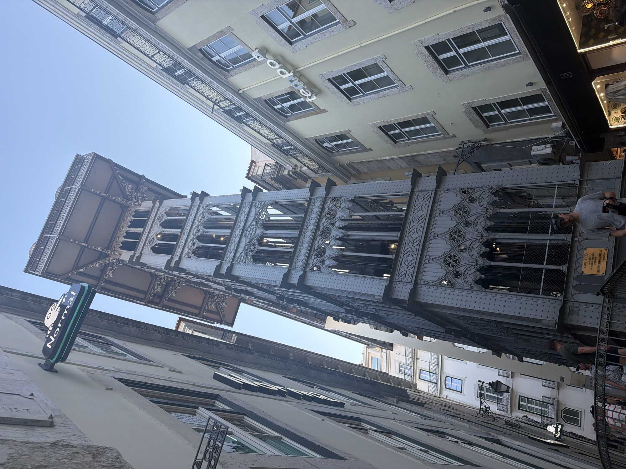 The image captures a view looking up between tall, narrow buildings in Lisbon, Portugal, bathed in the bright light of what appears to be daytime. The architecture suggests a historical European city with a mix of ornate ironwork and more traditional building facades.
Dominating the scene is the Santa Justa Lift, an impressive wrought-iron elevator tower with intricate gothic-inspired detailing. Its dark metal structure contrasts with the lighter colored buildings flanking it. One building to the left features numerous windows with stone surrounds and a sign that reads "Normal" in a modern, stylized font. Another building to the right shows a similar architectural style with multiple windows and a sign with some text that is partially visible. In the upper part of the image, more windows and balconies are visible on buildings that recede into the sky.
A few people are visible. One person is walking on a walkway of the Santa Justa Lift, appearing to be a tourist. Another person, wearing a grey shirt and a cap, is looking towards the camera from a balcony or walkway, partially obscured by the lift's structure.
The perspective is skewed, emphasizing the height and narrowness of the urban canyon. The clear blue sky is visible in the upper portion of the frame. The overall impression is one of urban exploration and appreciation for historical architecture in a bustling city.