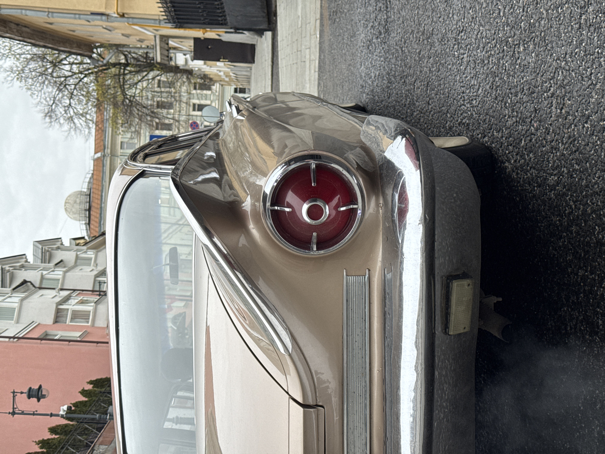 The image shows the rear of a vintage car, likely taken on a street in Moscow, Russia. The car is painted in a tan or beige color with prominent chrome trim around the taillight and rear bumper. The taillight is round with a red lens and a chrome cross pattern. The car's exhaust pipe is visible, emitting fumes. The background features a city street with buildings of varying heights and architectural styles, typical of older European or Russian cities. There is a streetlight and some trees in the backdrop. The road appears wet.