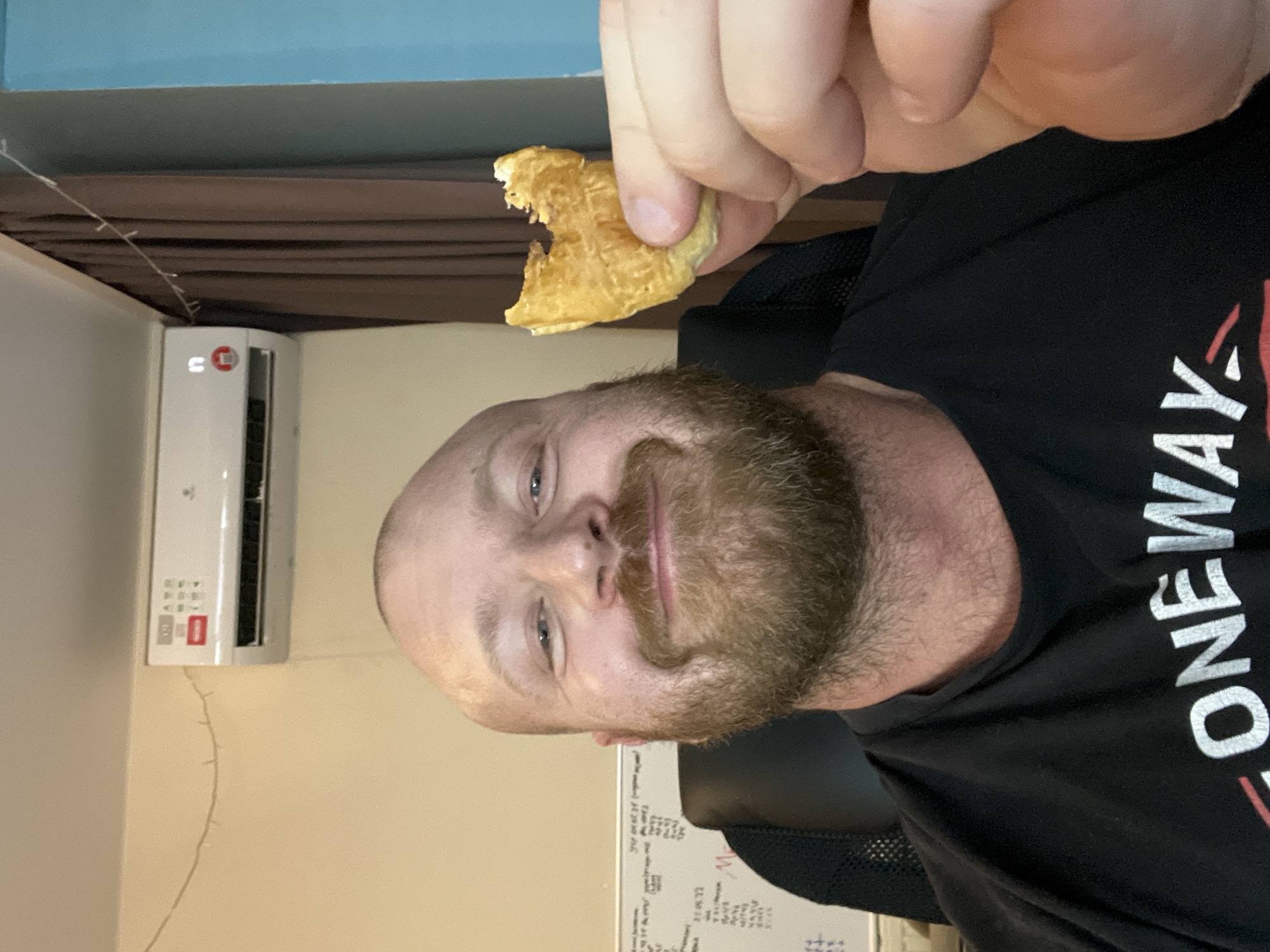 A selfie depicts a man with a shaved head and a red beard. He is holding a small, partially eaten pastry in his right hand. He is wearing a black t-shirt with white text that reads "ONEWAY." An air conditioning unit is mounted on the wall to the left of the man. A strand of lights is tacked to the wall, running from the air conditioner downwards. A dry erase board with writing is visible beneath the air conditioner. The background consists of a tan wall, a blue area above it, and a brown curtain. No specific city information is discernible.