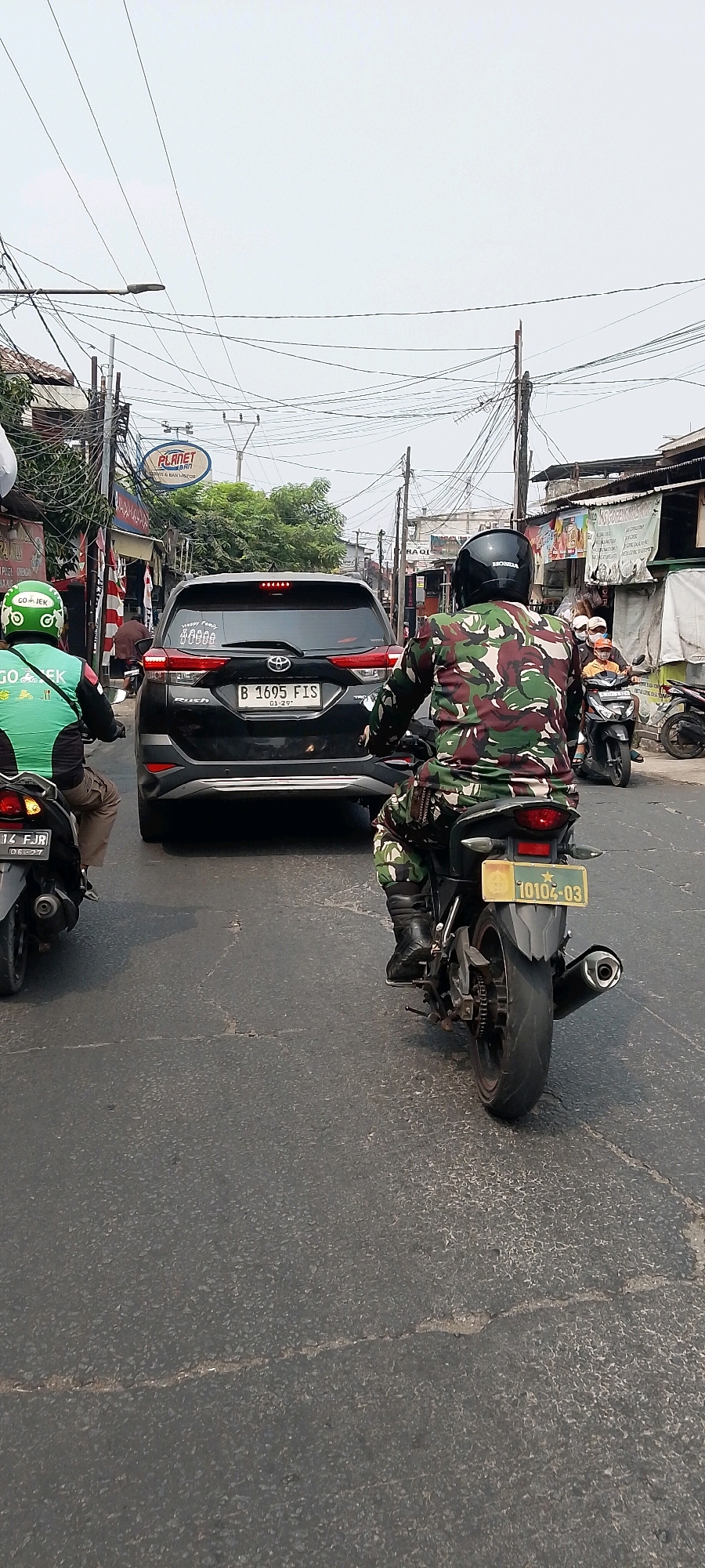 This is a street-level, eye-level, medium shot photograph taken in Bekasi, Indonesia, likely during the daytime given the bright sky and visible shadows. The scene depicts a traffic jam on a paved road lined with buildings and overhead power lines.
In the foreground, there are two motorcycles. The motorcycle on the left is partially visible and ridden by a person wearing a green Gojek uniform and helmet. The motorcycle on the right is ridden by an individual wearing a camouflage military uniform and a black helmet. The motorcycle has a yellow license plate with the number "10102-03".
In the midground, a black Toyota Rush SUV is stopped in traffic. Its license plate reads "B 1695 FIS". A sticker on the rear windshield appears to show a family silhouette with text above it. Further behind the SUV, more motorcycles and vehicles are visible, indicating heavy traffic.
The background shows a row of buildings with various signs, suggesting a commercial or residential area. One sign prominently displays "PLANET BRI". The sky is overcast and a complex network of power lines crisscrosses the scene above the street. The overall environment suggests a busy urban street in Indonesia.