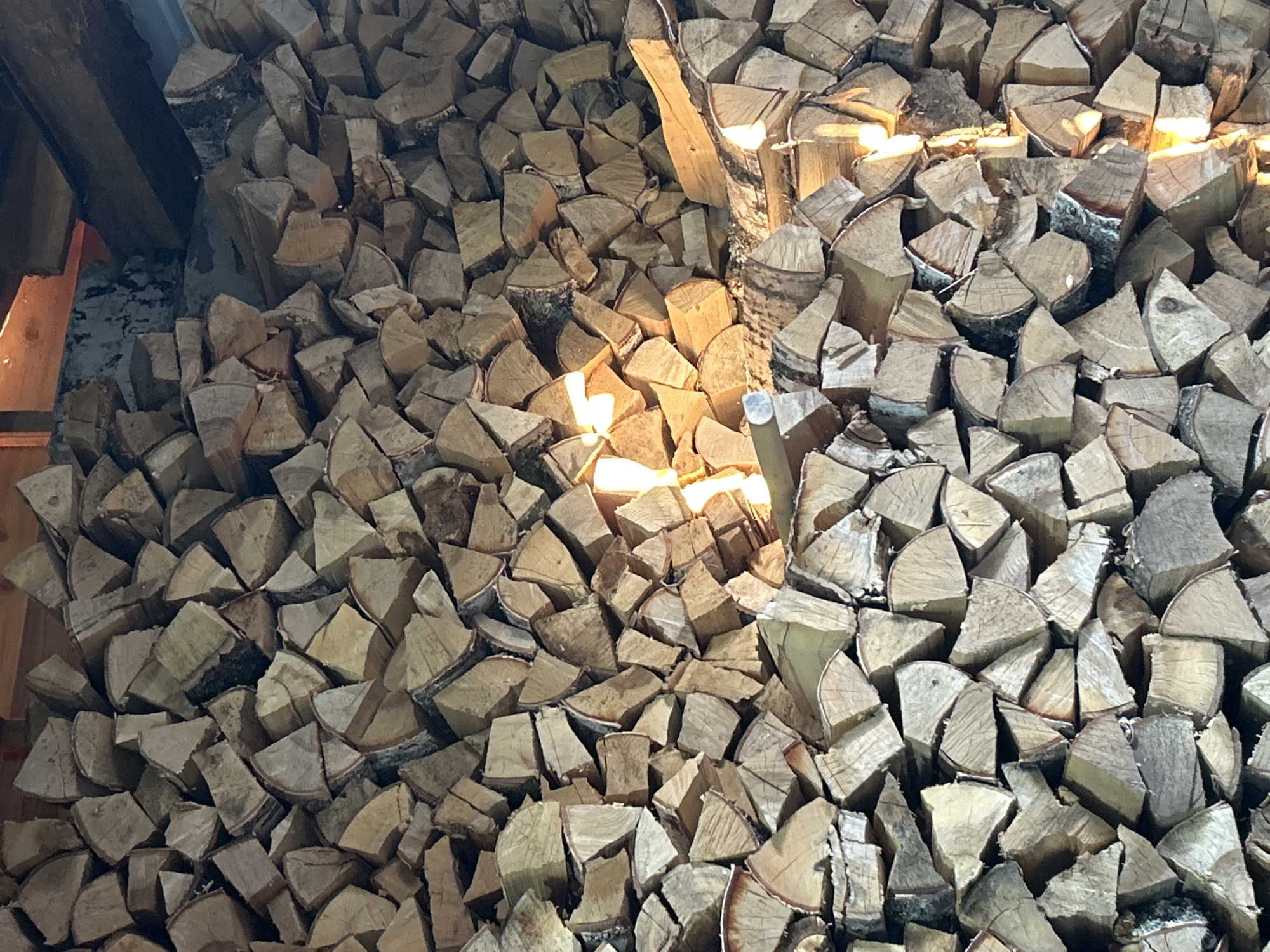 The image shows a densely stacked pile of chopped firewood. The wood appears to be seasoned and is of various sizes, predominantly in split log sections. The lighting suggests an indoor setting, possibly a shed or storage area, with warm light casting shadows and highlighting the texture of the wood. In the upper background, wooden beams are visible, indicating a roof structure. There are no people or animals in the image. The scene is static and focuses solely on the stacked firewood. The specific location in Chaikovsky, Russia, cannot be determined from the visual information provided in the image.