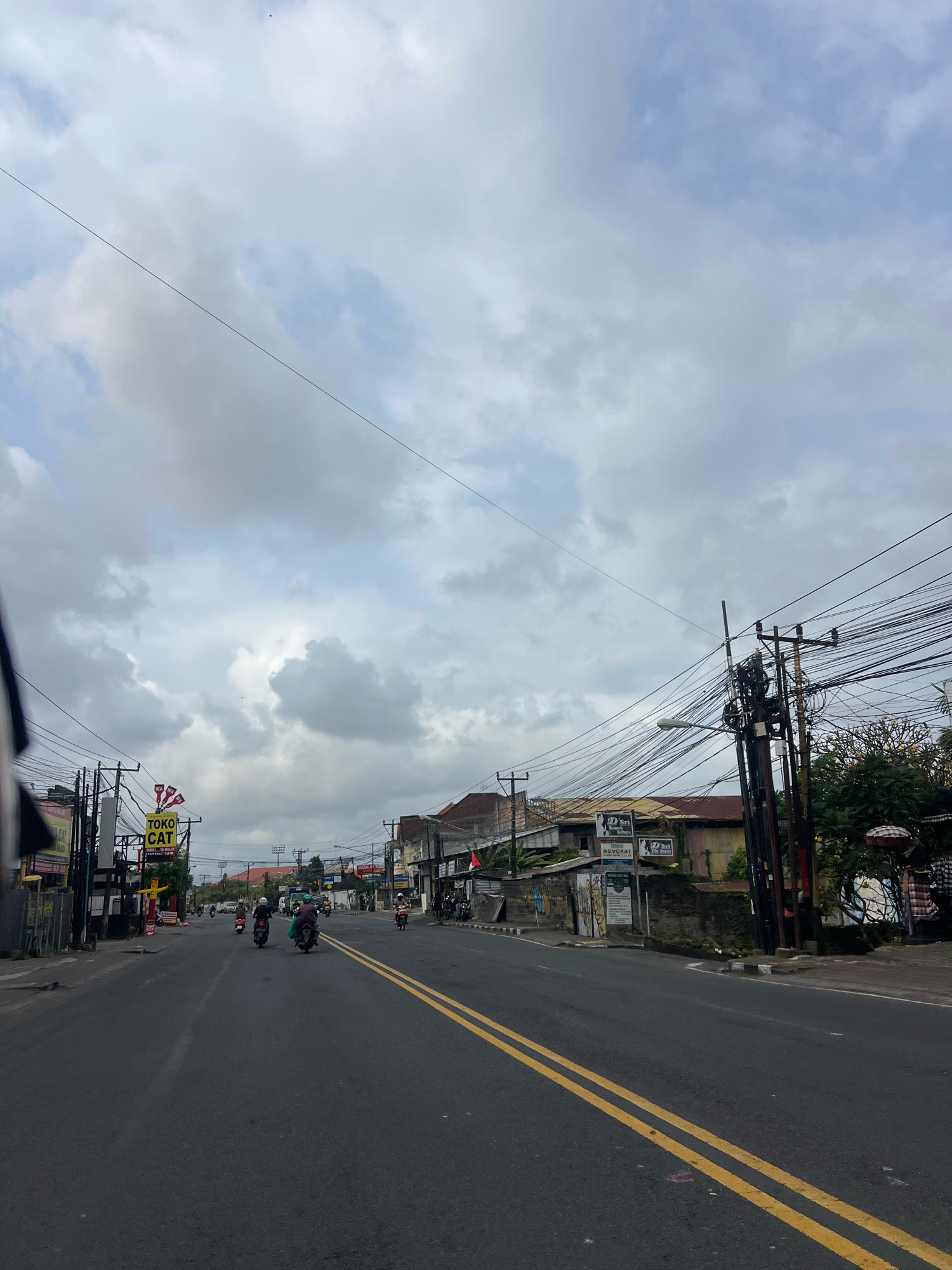 The image shows a street scene in Denpasar, Indonesia, likely on a cloudy day. A paved road with double yellow lines runs through the center of the frame, with several motorcycles traveling in both directions. Along the sides of the road are various buildings, some appearing to be shops or small businesses, with visible signage. One prominent sign reads "TOKO CAT" (Paint Shop) with a cartoon figure. Utility poles with a dense network of wires are visible on both sides of the street, along with streetlights. The architecture of the buildings suggests a tropical or Southeast Asian setting. In the background, more buildings and trees can be seen. The sky is filled with grey and white clouds.