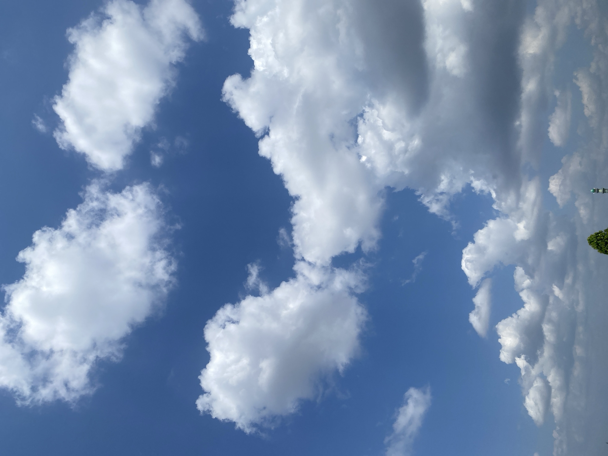 The image displays a wide expanse of a bright blue sky filled with fluffy white cumulus clouds. Some clouds are dense and billowy, while others are wispy and scattered. Towards the bottom of the frame, a sliver of land is visible, featuring a lone green tree on the left and a tall, slender white tower with a green cap on the right. This tower appears to be a minaret, suggesting the presence of a mosque. The overall scene depicts a clear, sunny day in Banjarmasin, Indonesia, likely captured from an elevated viewpoint, offering a panoramic sky view with a hint of the city's architecture.