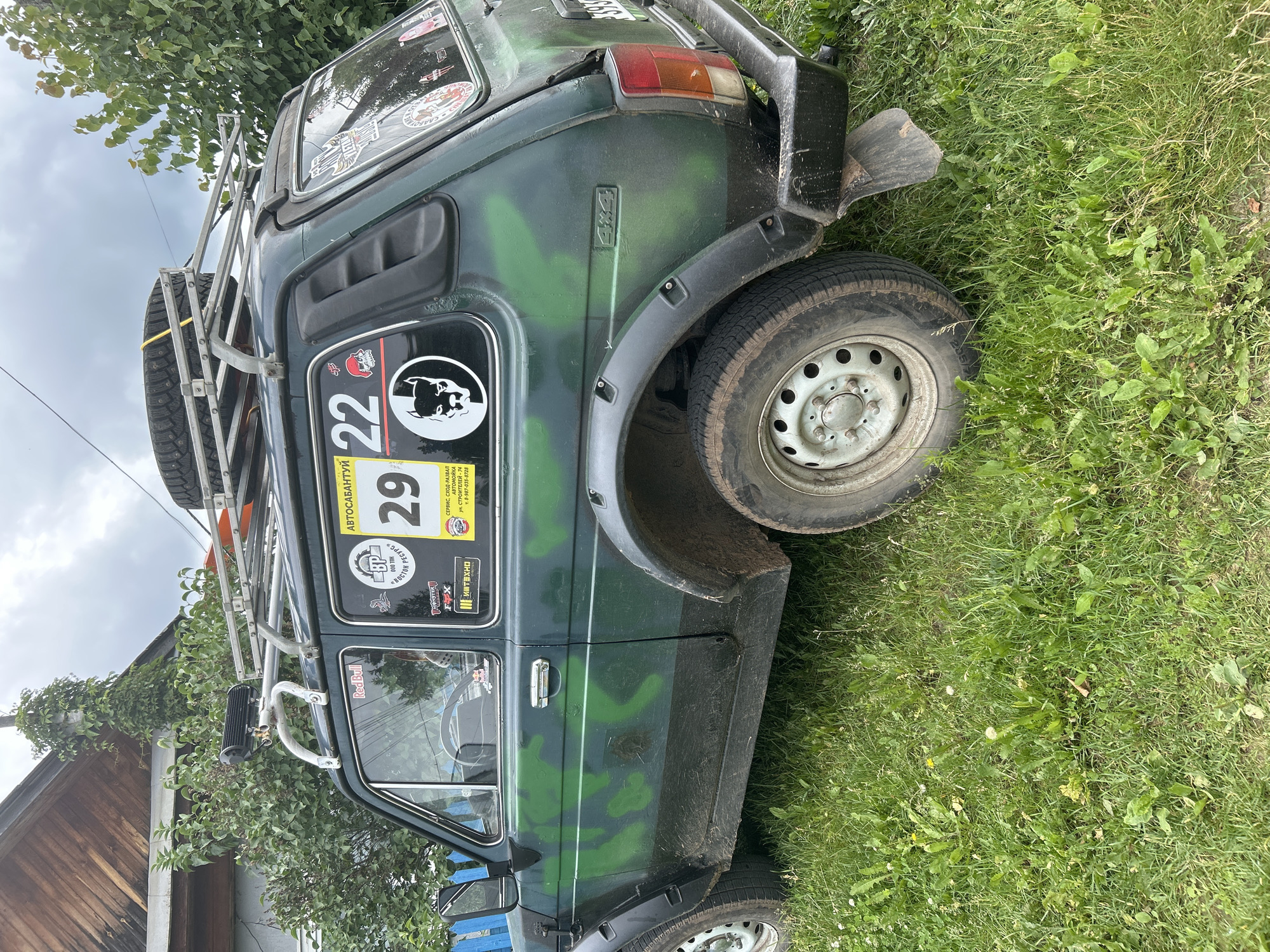 The image shows a dark green, camouflage-painted off-road vehicle parked on a grassy area next to a wooden building and some trees. The vehicle appears to be a Lada Niva, identifiable by its distinctive shape and the "4x4" badging. It is equipped with a roof rack holding a spare tire. Numerous stickers are affixed to the vehicle's windows and body, including logos for "Red Bull," "Monster Energy," "Fox," and what appear to be event or rally stickers with Cyrillic text. One prominent sticker displays the number "29" and another "22". A black shovel is leaning against the rear of the vehicle, partially obscured by the grass. The scene suggests an outdoor setting, likely in a rural or semi-rural location, with the sky appearing overcast. The overall impression is of a vehicle prepared for or used in off-road adventures or competitions. The visible text includes event names like "АВТОСАБАНТУЙ" (Autosabantuy, likely an event name) and service information in Russian for "СЕРВИС, СХОД-РАЗВАЛ АВТОМОЙКА" (Service, Wheel Alignment, Car Wash) with a phone number and street name.