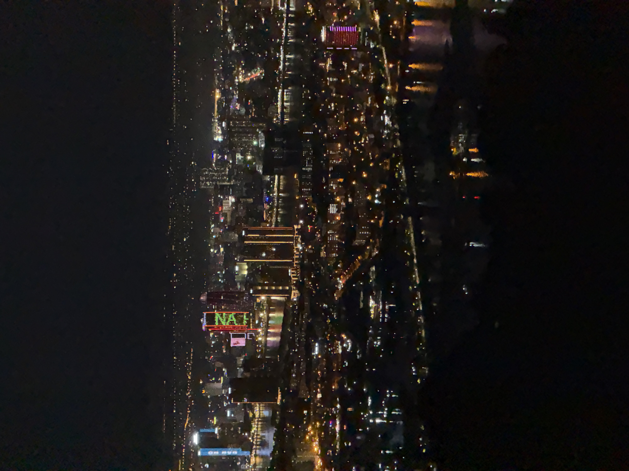 This is a nighttime aerial view of a cityscape in Da Nang, Vietnam. The image is dominated by darkness, with the city lights providing the main illumination. The scene depicts a dense urban environment with numerous buildings of varying heights, many of which are illuminated from within, casting a warm glow. A river or body of water runs through the city, reflecting the lights and creating streaks of color on its surface.
A prominent feature in the center of the image is a large building with a brightly lit sign displaying the text "NAH" in green and red capital letters. This sign is likely a prominent landmark or advertisement within the city. Other buildings also feature illuminated windows and architectural lighting. The overall impression is one of a bustling urban area at night, with the city alive with activity even after dark. The time of day is clearly nighttime, and there are no indicators of specific weather conditions. The location is identified as Da Nang, Vietnam, which is consistent with the dense urban landscape and the presence of a significant river or coastline. The image evokes a sense of urban energy and nighttime vibrancy.