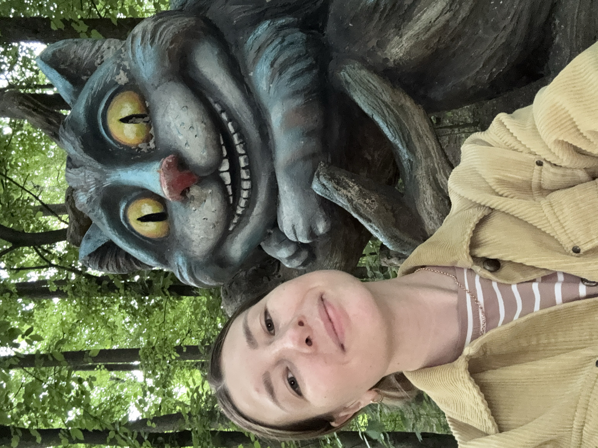 A young woman is captured in a selfie with a large, whimsical sculpture of the Cheshire Cat from Alice in Wonderland. The woman, with light brown hair and a subtle smile, looks directly at the camera. She is wearing a light yellow corduroy jacket over a striped top with beige and white or light pink vertical stripes, and a delicate gold chain is visible around her neck.
The prominent sculpture of the Cheshire Cat features large, glowing yellow eyes with dark slits, a wide, mischievous grin exposing many teeth, and a reddish nose. The cat's fur is depicted in shades of gray and blue, with visible texture and some weathering. Its body and paws appear to be integrated with or carved from dark, gnarled tree-like structures, suggesting it is part of a natural or themed outdoor setting.
The background is filled with lush green foliage, indicating an outdoor location like a park or garden, consistent with a public art installation in Moscow, Russia. The bright, diffused natural light suggests it is daytime. The image conveys a cheerful and playful mood, capturing an interaction with a fantastical character in a pleasant outdoor environment. No text is visible in the image.