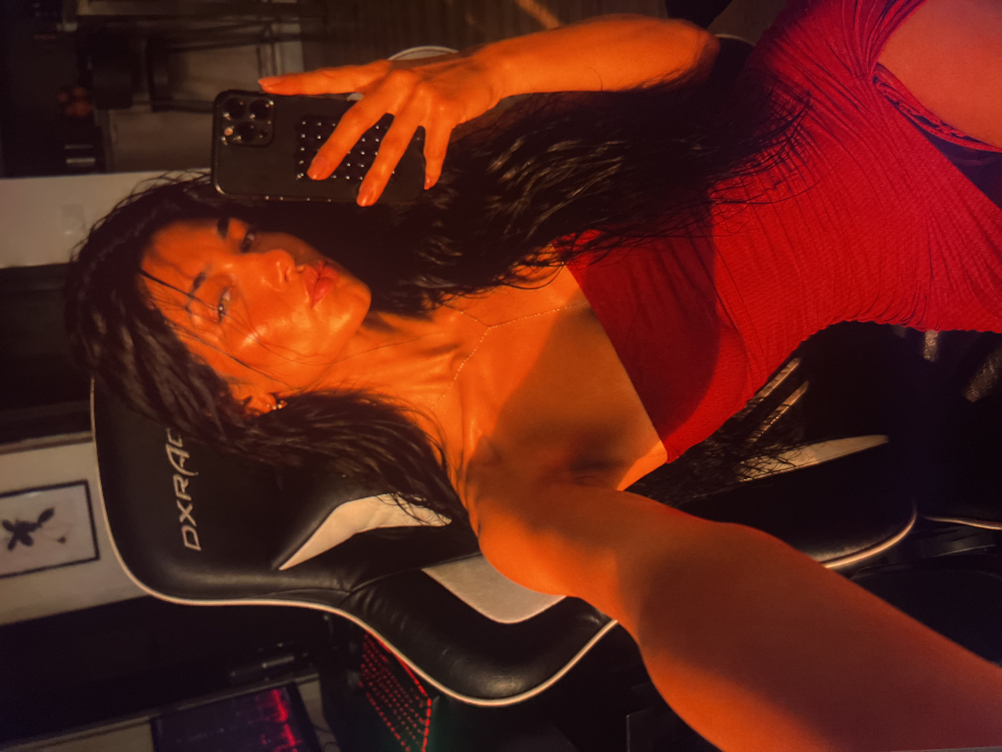 In an indoor setting, a woman with long, dark, wet hair takes a selfie with a smartphone. She is seated in a black and white gaming chair with "DXRACER" branding visible on the headrest. The woman is wearing a red strapless dress with a thigh-high slit. Her right arm is extended, holding the phone, while her left arm is bent at the elbow, her hand resting near her chest. The lighting in the scene is warm and orange-toned, casting strong highlights on her skin. In the background, a framed picture with a black abstract design is visible on the wall, and a gaming computer setup with illuminated red keys is present to the left. Further in the background, a kitchen counter with what appear to be fruit and stools can be seen. The overall impression is an intimate, self-taken photograph in a personal space.