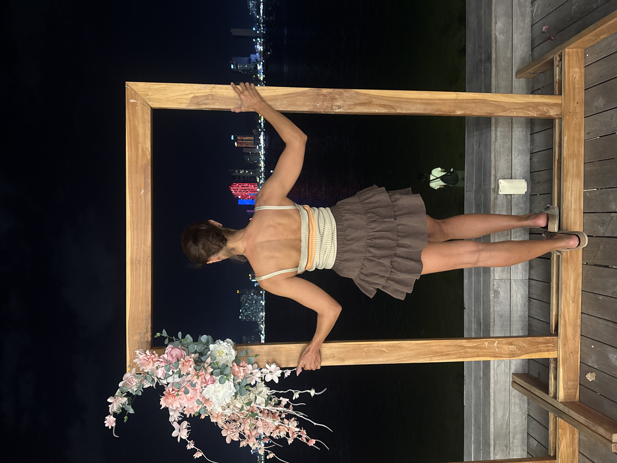 The image captures a nighttime scene in the city of Nha Trang, Vietnam. A woman is standing within a large wooden frame, her back to the camera. She is wearing a light-colored, striped crop top and a brown ruffled skirt. Her hands are resting on the wooden frame, one on each side. To the left of the frame, a floral arrangement with pink and white flowers is attached. The background features a body of water with a cityscape illuminated by city lights and buildings in the distance. A person is visible in the water behind the woman, seemingly swimming or wading. The foreground consists of a wooden deck.