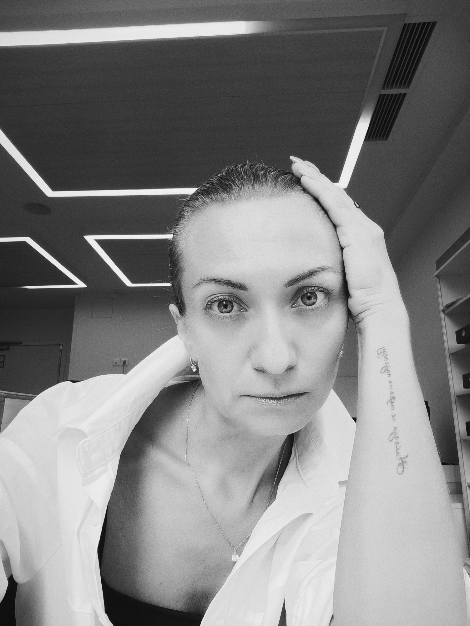 A black and white, medium shot selfie shows a woman with her right hand on her head and her left arm resting on her chest. She is wearing a white button-down shirt, unbuttoned, revealing a dark bra underneath. A delicate necklace with a pendant is visible. She has dark hair pulled back and appears to be looking directly at the camera with a serious expression. A tattoo is visible on her left forearm. The background features modern ceiling lighting, ventilation grates, and a blurred office-like environment with shelves in the background. The scene is indoors, likely in an office in Moscow, Russia.