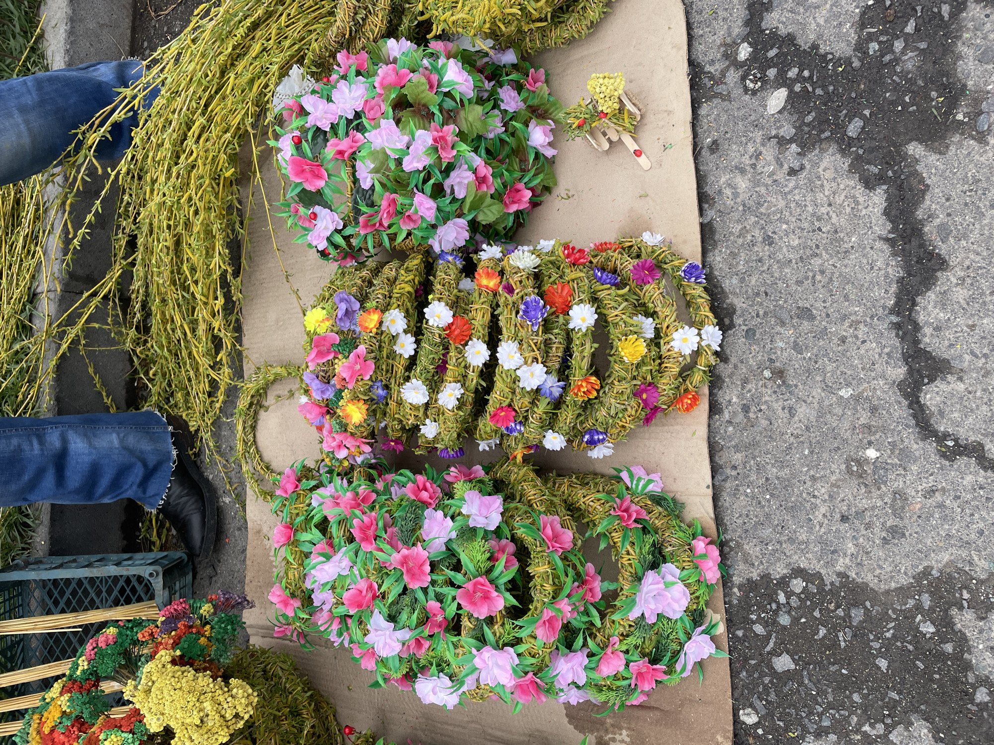 The image shows a selection of wreaths and floral arrangements displayed on a piece of cardboard on a Yerevan, Armenia street. There are four visible wreaths made of bound branches and adorned with artificial flowers. Two wreaths have pink and light pink flowers with green leaves. One wreath has small white, orange, yellow, and purple flowers. A fourth partially visible wreath is composed of yellow and red flowers. To the left of the wreaths, there is a bundle of long, thin yellow branches. To the right of the wreaths, there is a small figure constructed from wood sticks and yellow flowers. The corner of a blue plastic crate is visible next to a person's legs in blue jeans and black shoes. The background is a gray paved street.