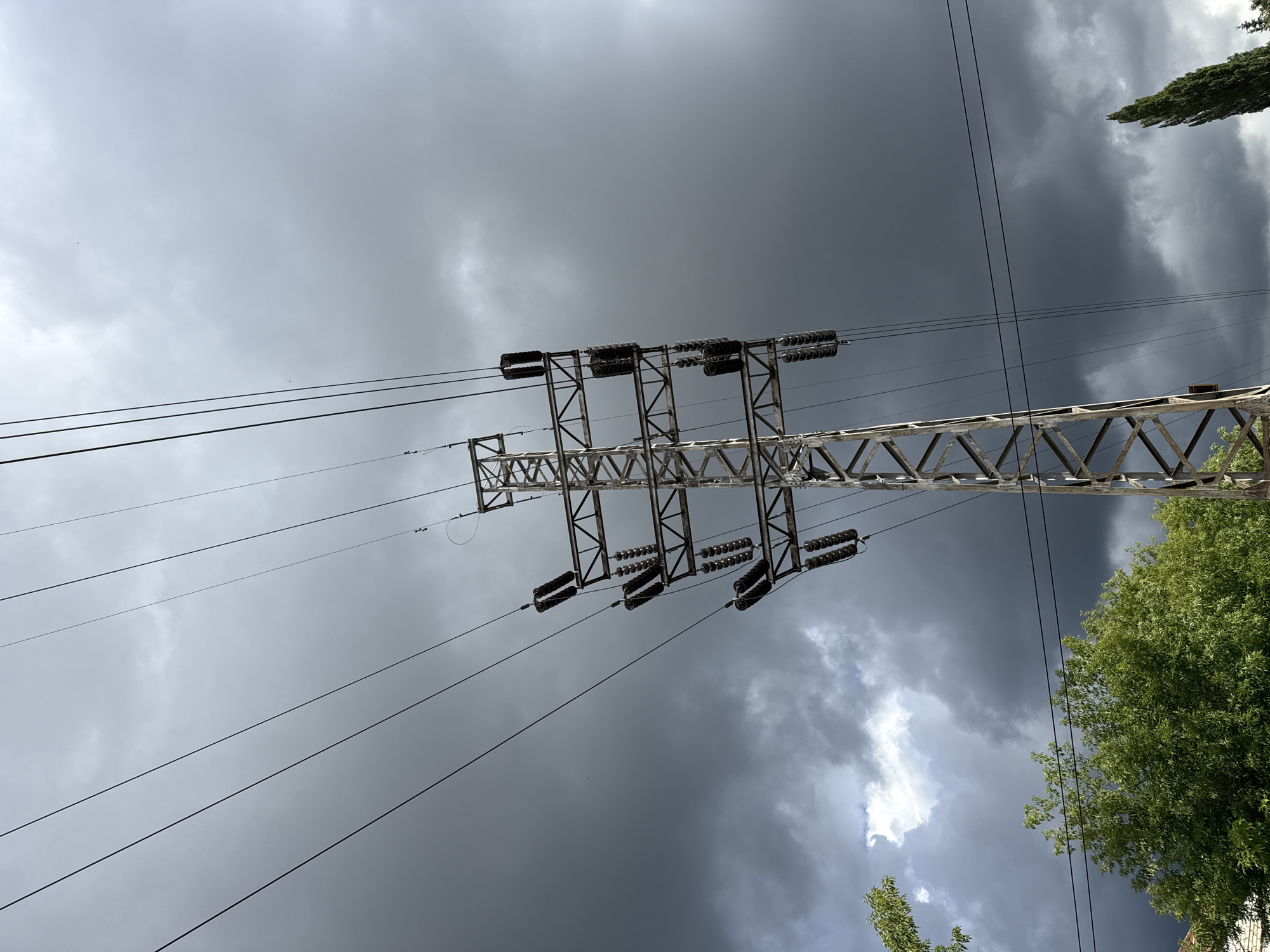 An outdoor, low-angle, vertical shot captures a metal electricity pylon with multiple sets of insulators holding power lines against a dark, cloudy sky. The pylon is a lattice tower structure made of metal beams. Several sets of dark-colored insulators hang from the horizontal arms of the pylon, with power lines strung between them. The sky is filled with heavy, gray storm clouds, with a patch of lighter sky visible towards the upper left. In the lower left corner, the tops of green trees and a small portion of a brick building are visible. On the right side, a tall, slender cypress tree stands against the dark sky. The image appears to be taken in an urban or semi-urban environment.