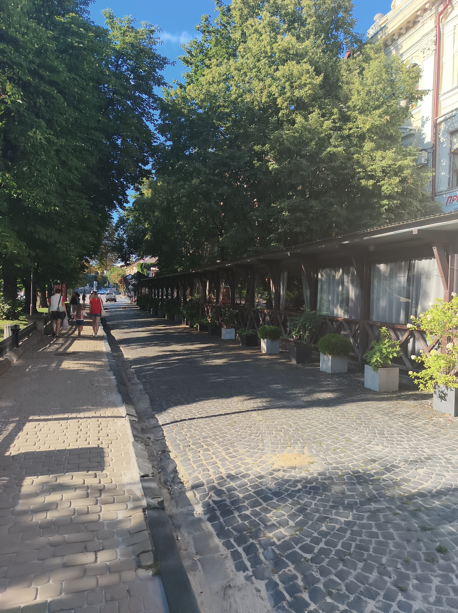 The image depicts a street scene in Ternopil, Ukraine, on a sunny day. On the left side of the street, a pedestrian walkway paved with light-colored bricks is visible. A low, dark railing separates the walkway from a grassy area beyond. Several individuals are walking along the brick path, including a group of four people in the foreground, two adults and two children. They are walking away from the camera.
The right side of the street features a cobblestone road. Adjacent to the cobblestones, a long, wooden, open-air structure with a roof provides shade. This structure appears to be an outdoor seating area for a cafe or restaurant, with tables and chairs likely situated within, though not explicitly visible. The structure is lined with potted plants.
The street is lined with mature trees, casting shadows on the walkway and road. In the background, a building with decorative architectural details and a sign with Cyrillic lettering is visible. The sky is blue with a few wispy clouds. The scene conveys a sense of urban life on a pleasant day.