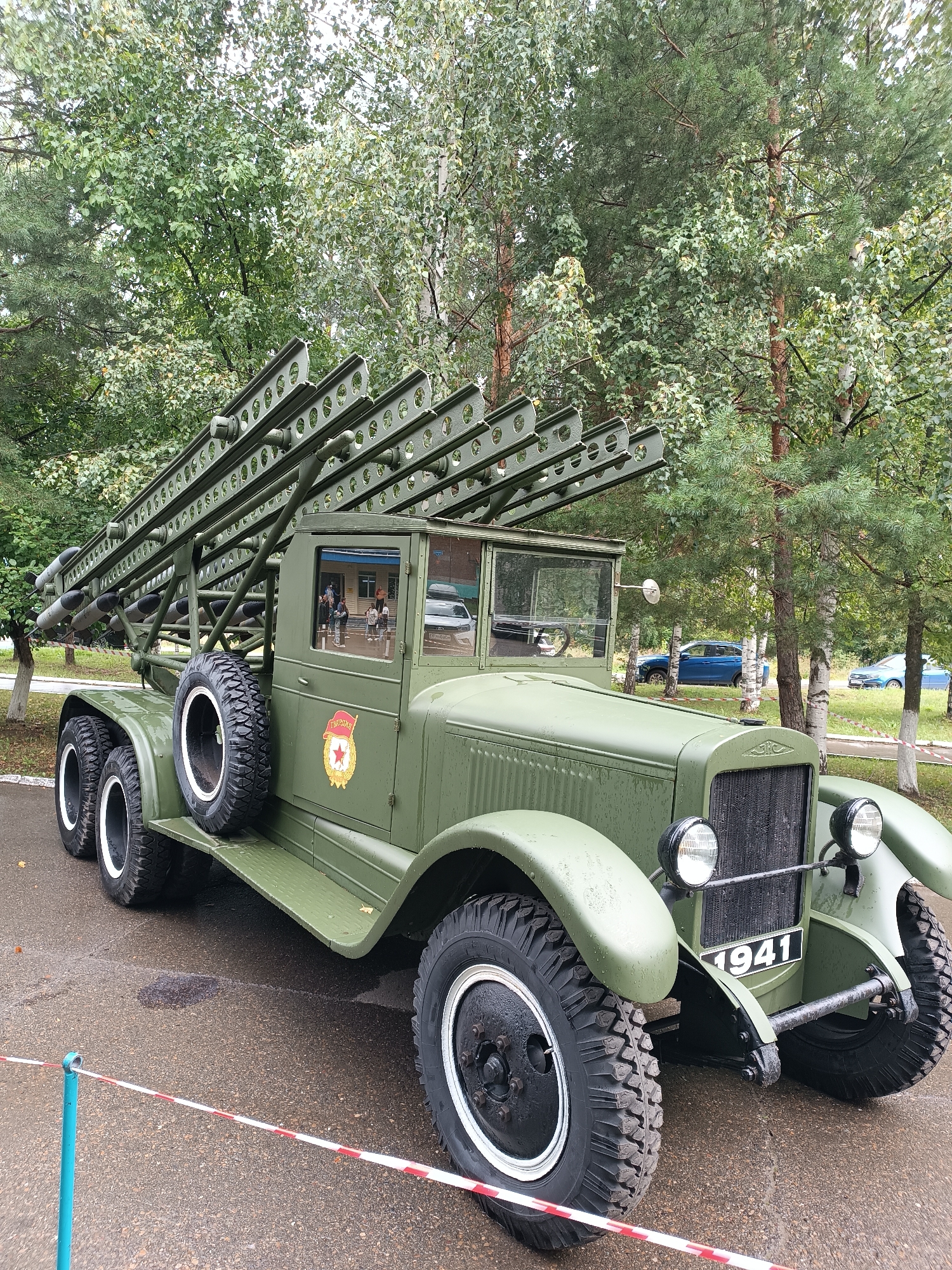 A military truck, painted olive drab, is parked on a wet asphalt surface in an outdoor setting. The truck is equipped with a rack of multiple rocket launchers mounted on its bed. The vehicle features large, heavy-duty tires, a prominent grille, and round headlights. A license plate on the front reads "1941". A red and white striped barrier tape is visible in the foreground, indicating a restricted area. In the background, a wooded area with a variety of trees, including birch trees, forms a natural backdrop. Several people are visible inside the truck's cabin, appearing to be observing the surroundings or interacting with each other. Behind the truck, other vehicles, including a blue car, are parked. The sky is overcast. The scene is likely a historical display or a museum exhibit, given the vintage military vehicle. The location is the city of Chaikovsky, Russia.