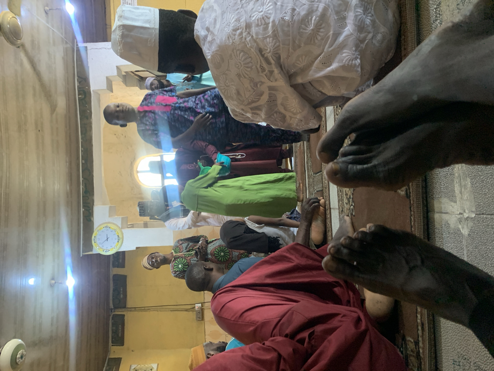 The image depicts an indoor scene, likely a mosque, in Lekki, Nigeria, with several individuals present. In the foreground, two bare feet are visible, suggesting the photographer is seated on the floor. The individuals in the background are mostly men and boys dressed in traditional Islamic attire. A man in a blue shirt and red robe is seated, facing away from the camera. To his right, a person in a green robe is standing. Further back, a young boy in a patterned shirt and pink accents is looking towards the camera. Other individuals are blurred or partially obscured.
The environment appears to be a place of worship, with wooden ceilings and yellow walls. A circular clock with a yellow frame is mounted on the wall, displaying the time. A ceiling fan is also visible. The floor is covered with patterned rugs. The lighting suggests it is daytime.