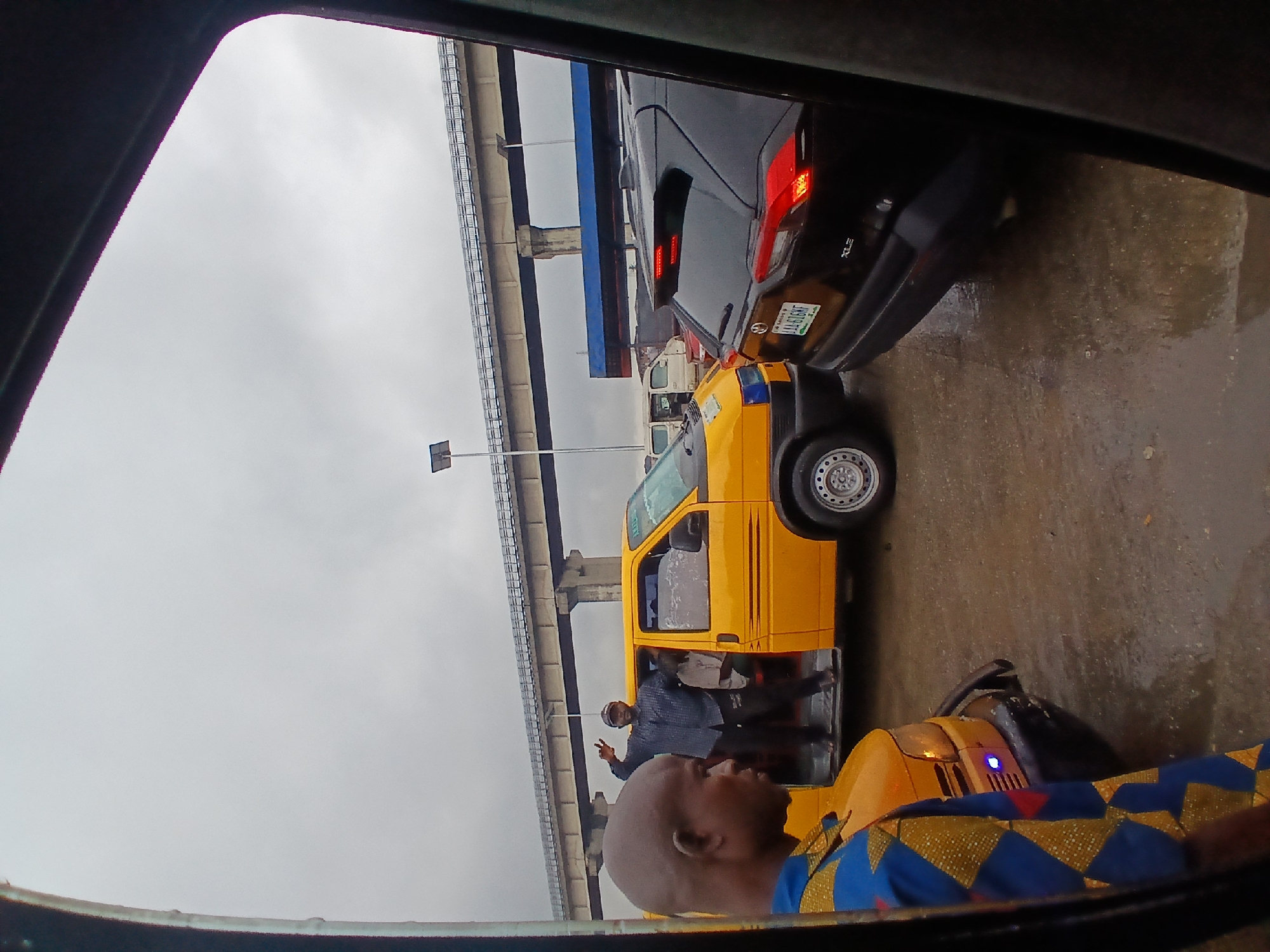 The image depicts a scene in a congested urban environment, likely Agani, Nigeria, characterized by a cloudy, overcast sky and wet asphalt road. In the foreground, a person is visible from the chest up, wearing a blue, yellow, and red patterned shirt. To their left, a yellow minibus is parked, its front passenger door open. A man stands in the doorway of the minibus, wearing a checkered blue shirt and dark pants, with his right arm raised in a gesture of greeting or acknowledgment. Another individual, partially visible, is seated inside the minibus.
Further into the scene, a black SUV is positioned behind the yellow minibus, its brake lights illuminated. A portion of the SUV's license plate is visible, showing alphanumeric characters. In the background, an elevated concrete structure, possibly an overpass or a bridge, spans across the scene, supported by concrete pillars. A lamppost with a solar panel is situated near the overpass. The overall atmosphere suggests a busy, possibly rainy day in a Nigerian city, with vehicles and infrastructure indicative of a developed urban area.