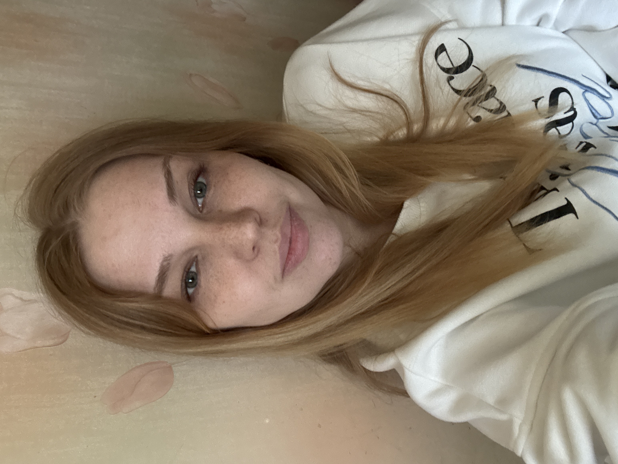A woman with freckles and auburn hair lies on a light brown surface next to a few pale pink objects that resemble petals. She is wearing a white sweatshirt with black and blue lettering. The background appears to be a wall. The location is not possible to determine from the image.