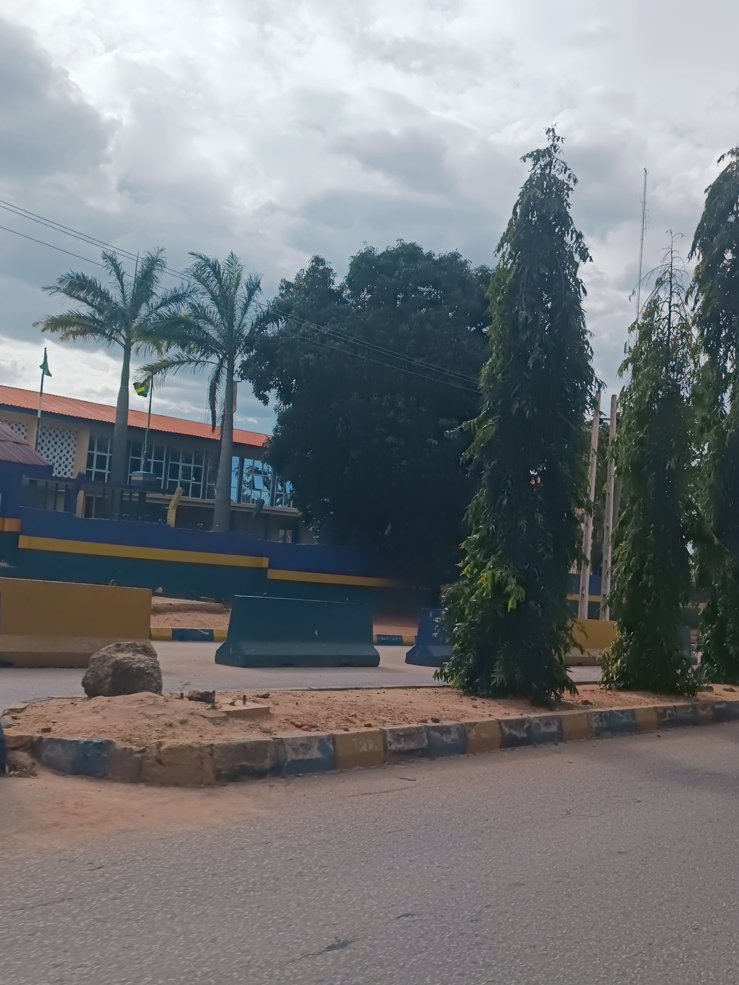 The image shows the exterior of a building, likely a government institution, located in Jos, Nigeria. In the foreground, there is a paved road and a concrete curb. Beyond the curb, a sandy area with scattered rocks and concrete barriers is visible. A large building with a red-tiled roof and a facade featuring white patterned sections and large blue-tinted windows stands behind these barriers. Two palm trees are positioned in front of the building, and two flags are flying from poles. The sky is overcast with gray clouds. Several tall, slender trees with dense foliage are present on the right side of the image, partially obscuring the view of the building. The overall impression is of an institutional or official compound on a cloudy day.