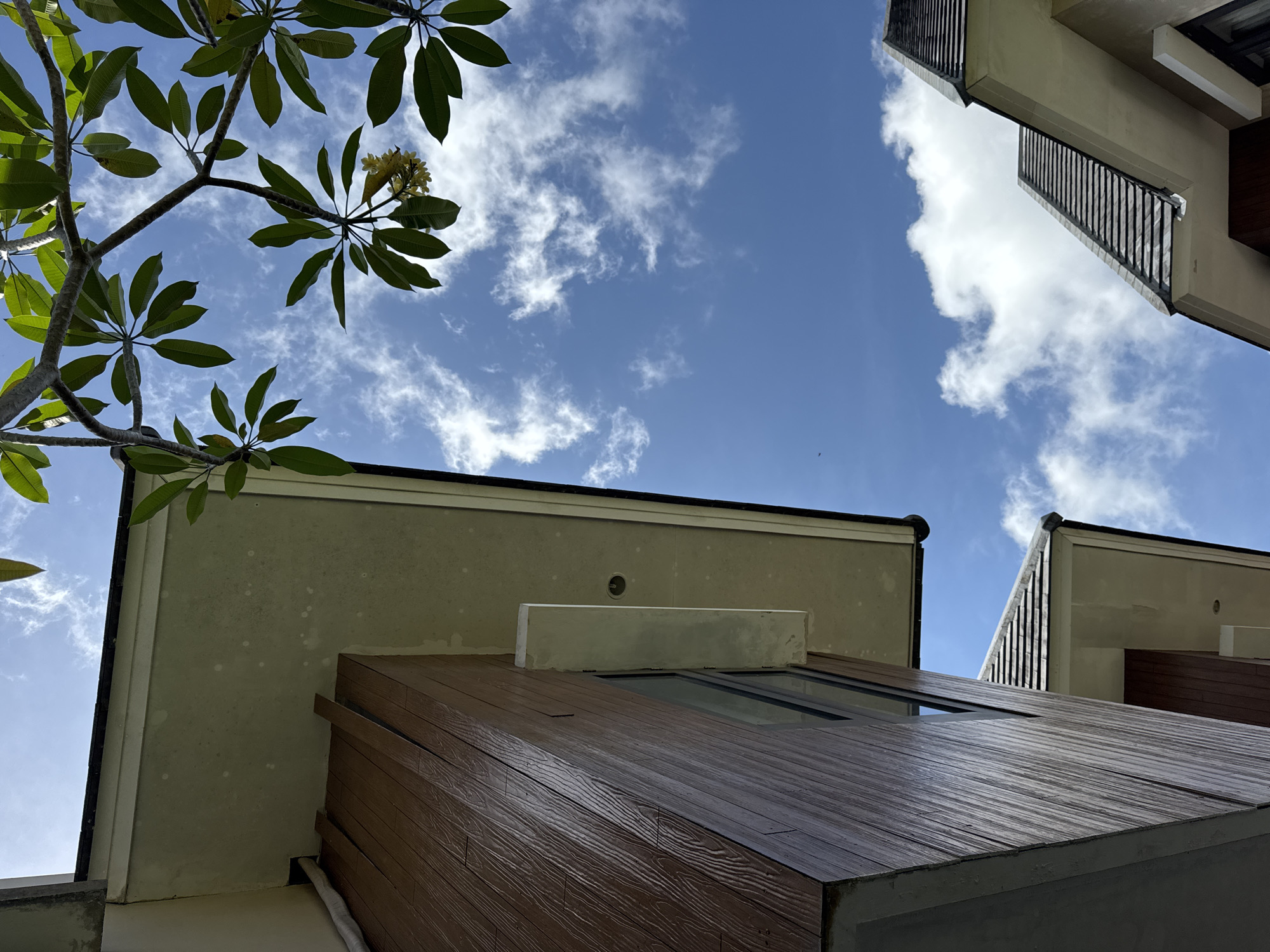 The image is taken from a low angle, looking up towards a blue sky with scattered white clouds. In the upper left, a tree branch with green leaves and yellow flowers partially obscures the sky. The lower portion of the frame is filled with architectural elements. There are multiple building facades in view, featuring a combination of light-colored stucco or concrete surfaces and brown wood-like cladding. One section has a small rectangular window. The location is Kuta Selatan, Indonesia.