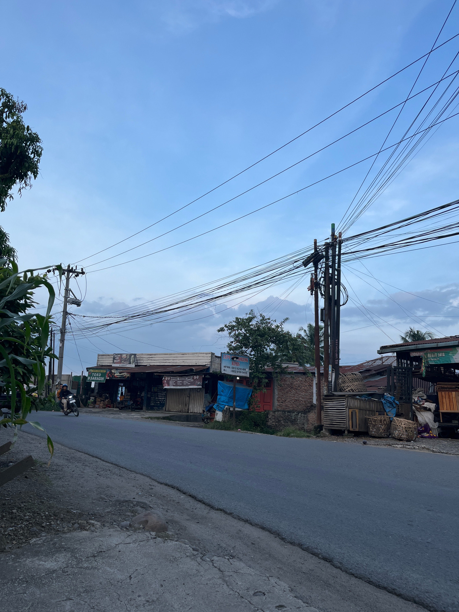 The image depicts a street scene in Patumbak Dua, Indonesia, characterized by a paved road running horizontally across the frame. On the right side of the road, several small shops and buildings are visible, some with open fronts displaying goods. In front of these establishments, there are various objects, including wooden crates, baskets, and covered items, suggesting commercial activity. A tall utility pole with a dense network of electrical wires dominates the upper right portion of the image. Vegetation, including trees and leaves, frames the left side of the scene. A motorcycle with a rider is traveling on the road, moving away from the viewer. The sky above is partly cloudy with a pale blue hue. The overall atmosphere suggests a moderately populated urban or semi-urban area during daylight hours.
