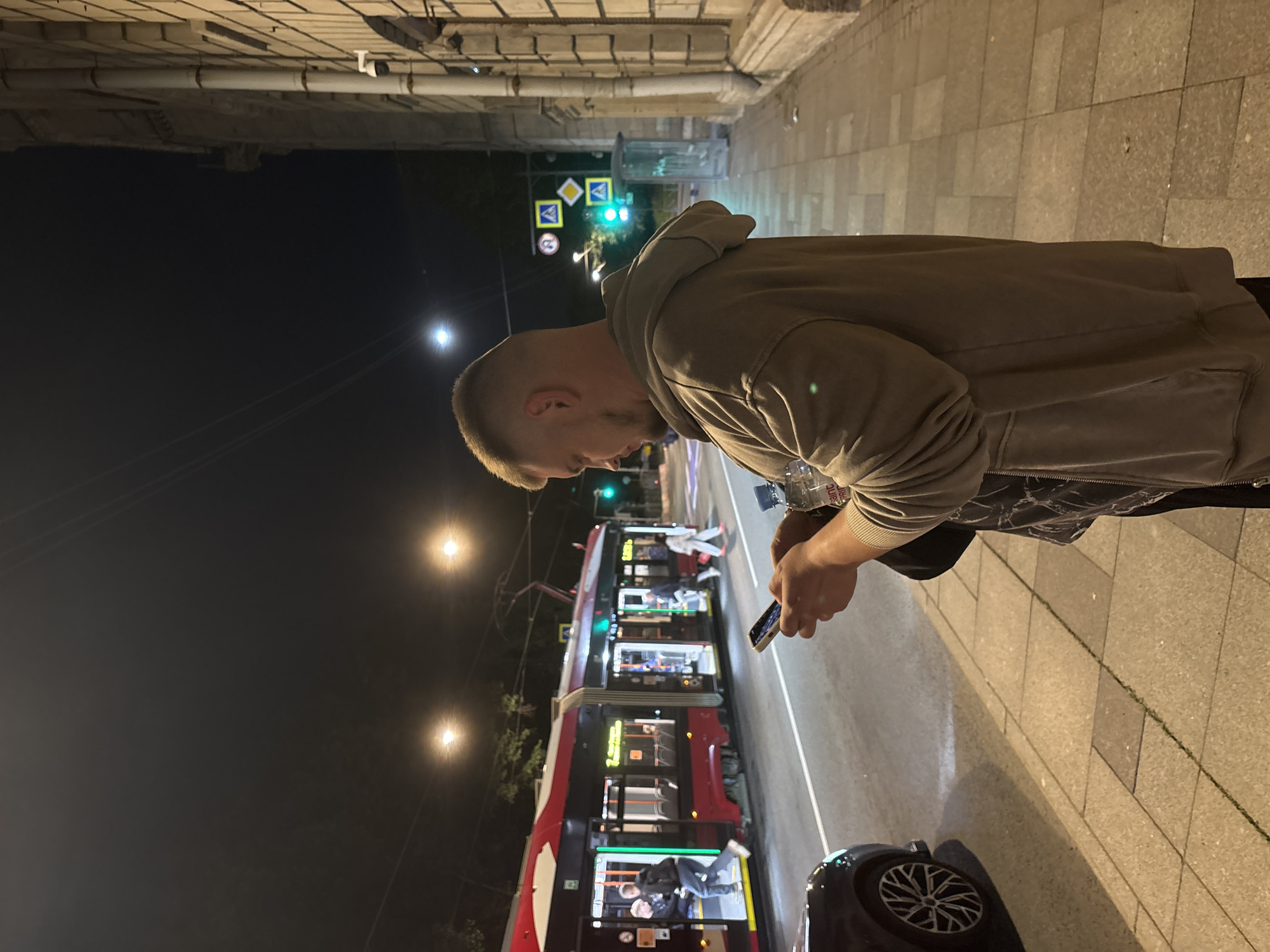 A man stands on a sidewalk at night, looking at his phone. He is wearing a tan hoodie and holding a water bottle. In the background, a red and black tram is stopped at a station, with people boarding and disembarking. Streetlights illuminate the scene, casting long shadows. The tram appears to be on a street in Saint Petersburg, Russia, indicated by the presence of a tram and the architectural style of the building on the right side of the image, which is characteristic of the city. Traffic lights and road signs are also visible.