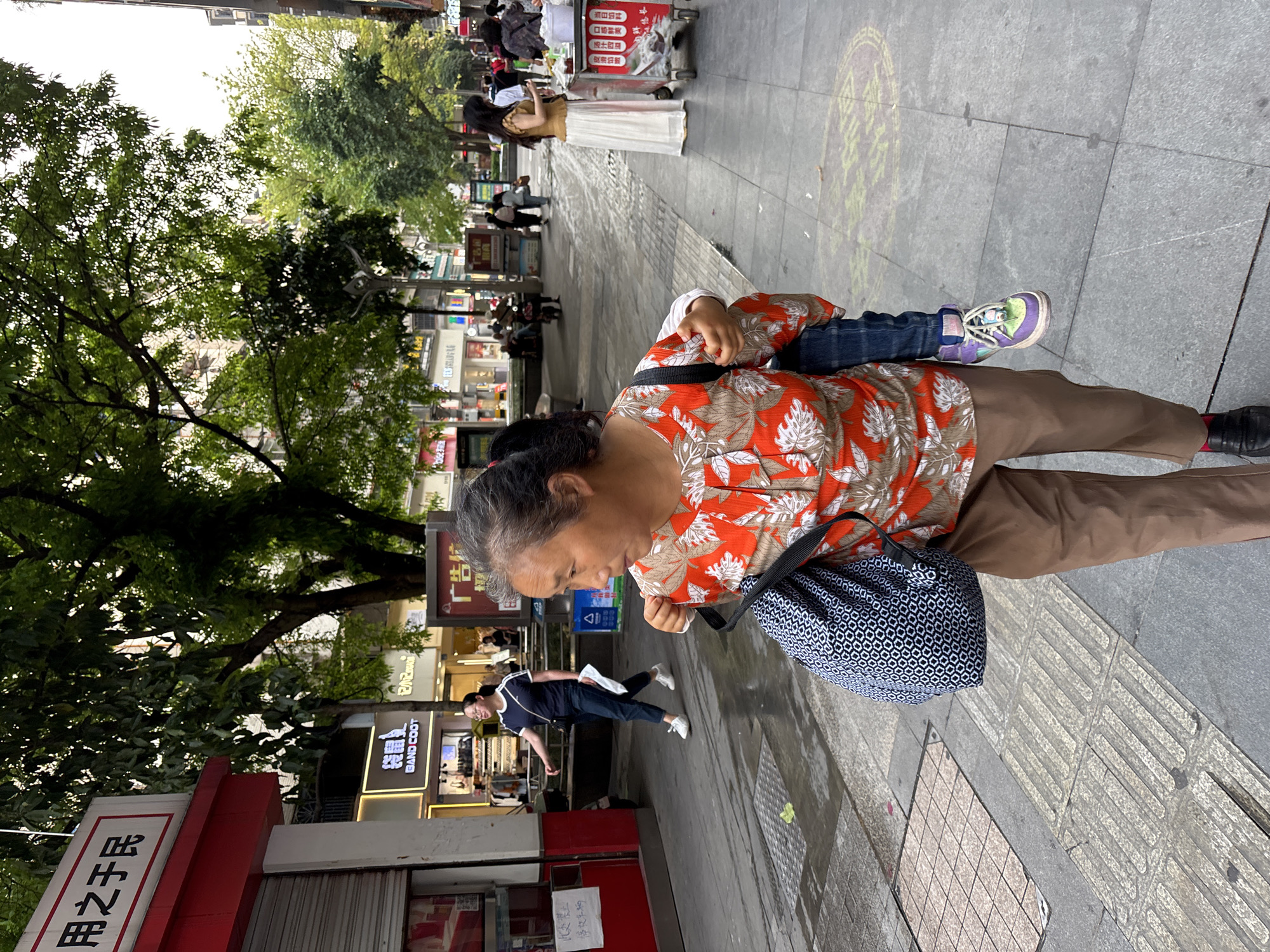 The image shows an elderly woman carrying a child on her back on a sidewalk in the city of Jiangjin, China. She wears an orange and white patterned shirt and brown pants. A blue and white patterned bag hangs from her shoulder. The child is wearing jeans and multi-colored sneakers. The street is lined with shops, trees, and pedestrians. Signs in Chinese script are visible on the storefronts. The words "BAND COOT" are visible on one storefront.