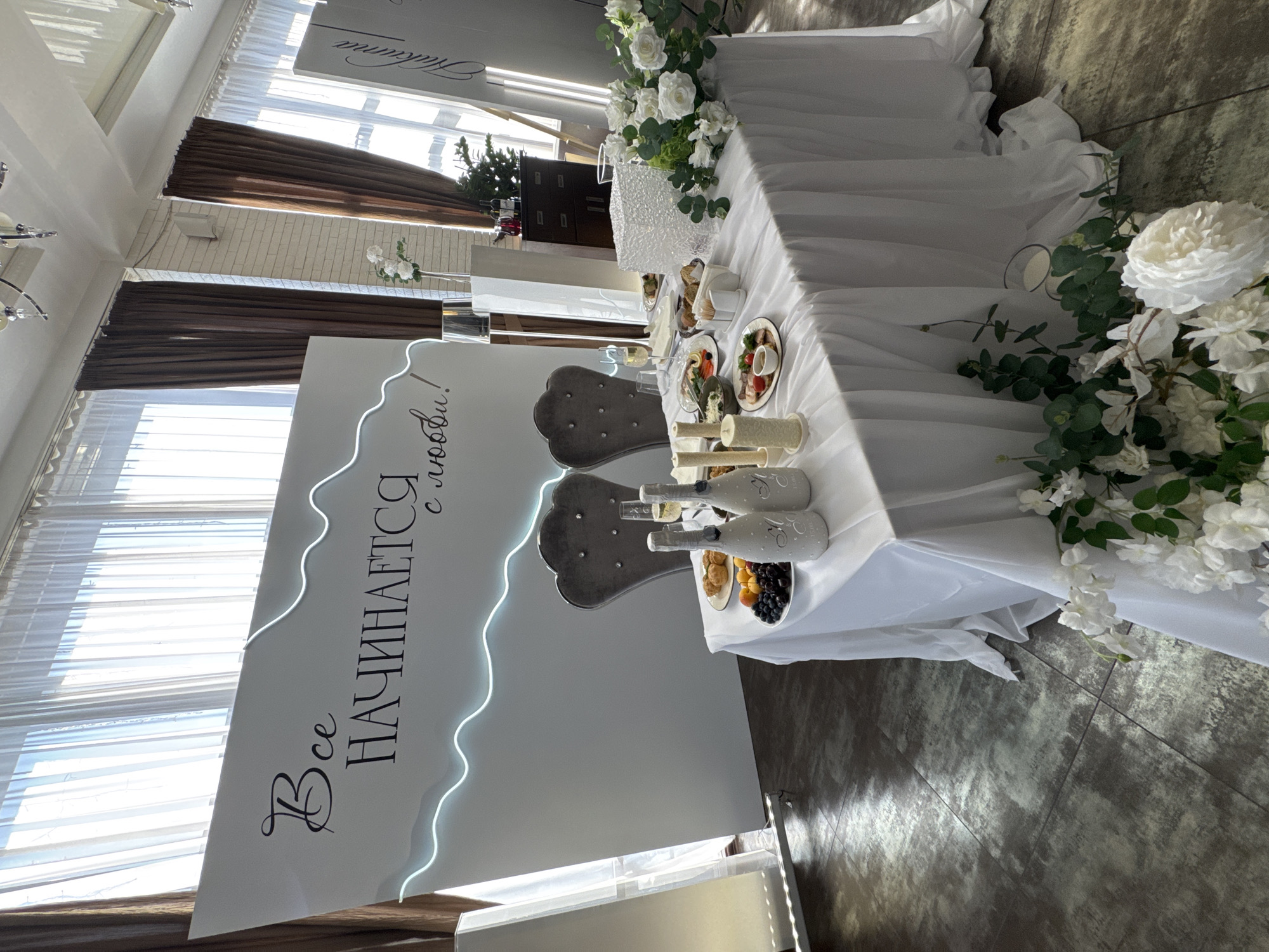 The image depicts a decorated wedding reception setup. A white rectangular backdrop features a Cyrillic inscription reading "Всё начинается с любви!" ("Everything begins with love!") in black text, accented by a wavy neon light strip. In front of the backdrop, a table is draped with a white tablecloth, laden with food, drinks, and floral arrangements. Two chairs with high, tufted backs are positioned at the table. On the table, there are two bottles of champagne, plates of food including fruit and pastries, candles, and a clear cube-shaped object possibly used as a centerpiece or light feature. A bouquet of white roses and greenery is arranged on the right side of the table. The floor appears to be tiled with a dark, textured pattern. The environment suggests an indoor event space, potentially a restaurant or banquet hall.