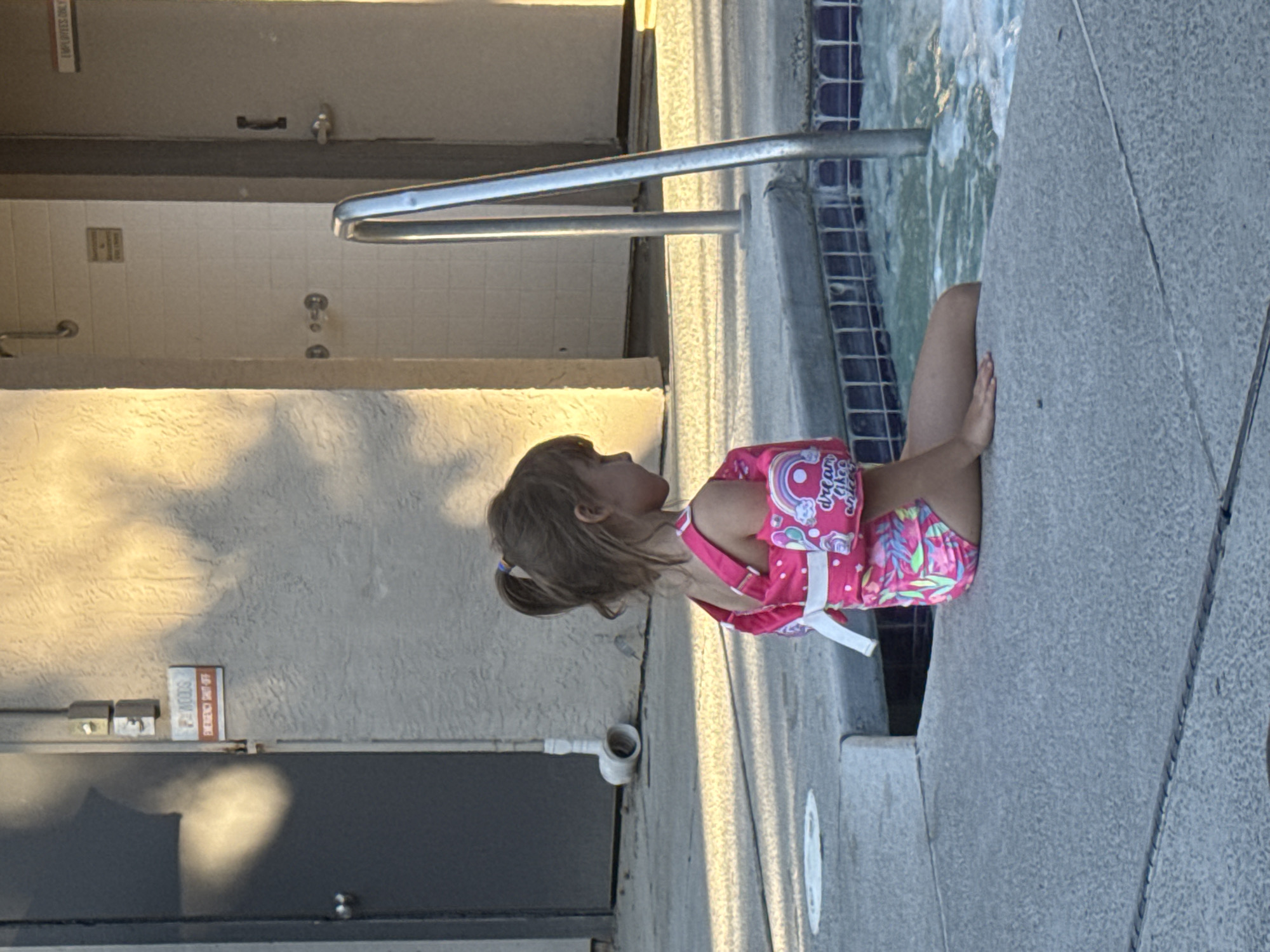 A young girl wearing a pink swimsuit with unicorn designs and arm floaties sits on the edge of a tiled swimming pool. The girl is positioned facing left in profile, with her right arm resting on her lap and her left arm slightly extended. She has brown hair pulled into a ponytail. The pool area appears to be outdoors, with a concrete deck surrounding the pool. In the background, there is a building with a textured wall, a gray door with a handle, and an "Employees Only" sign. There is also a metal handrail entering the pool. The lighting suggests it is late afternoon, casting shadows on the wall. A red and white sign on the wall reads "P-WOODS EMERGENCY SHUT-OFF". A white PVC pipe extends from the wall near a door.