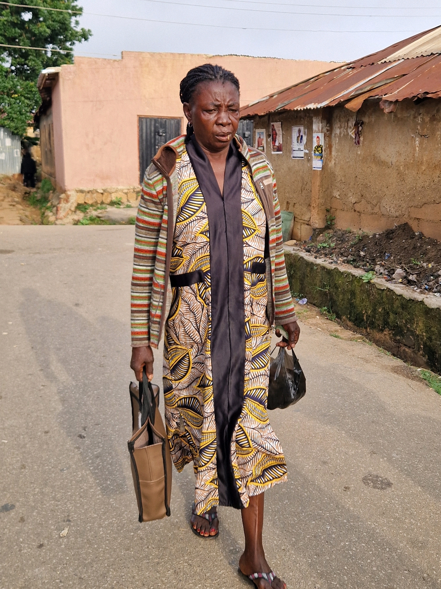 A middle-aged Black woman with dark skin and braided hair walks towards the camera on an asphalt road in Kabong, Nigeria. She is wearing a long, dark dress over a patterned robe with yellow, white, and brown motifs and a striped cardigan. The woman carries a brown tote bag in her left hand and a black plastic bag in her right. The background shows a light pink building with a metal roof, a brown and beige building with posters on its wall, and a concrete drainage ditch filled with debris. The scene appears to be a residential area.