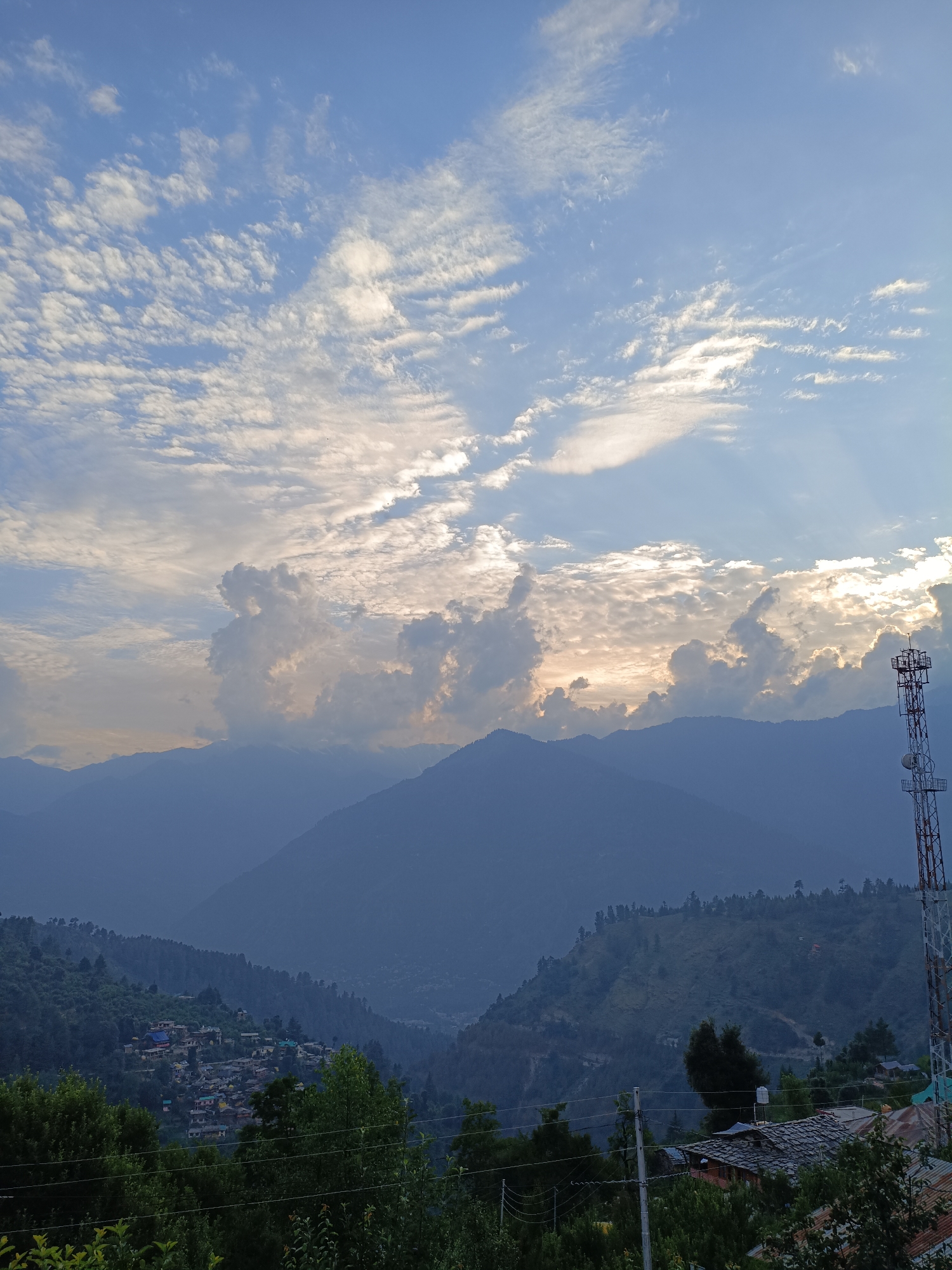 The image captures a panoramic view of a mountainous landscape in Damadi, India, likely at dusk. The sky is a blend of blue and white, with scattered clouds, some of which are illuminated by the setting sun, casting a warm glow. In the foreground, lush green trees partially obscure a cluster of houses nestled on the slopes of the hills. Further in the distance, more mountains are visible, fading into a hazy blue silhouette. A prominent cell tower stands to the right of the frame, its structure silhouetted against the sky. The overall scene is tranquil and serene, showcasing the natural beauty of the region.